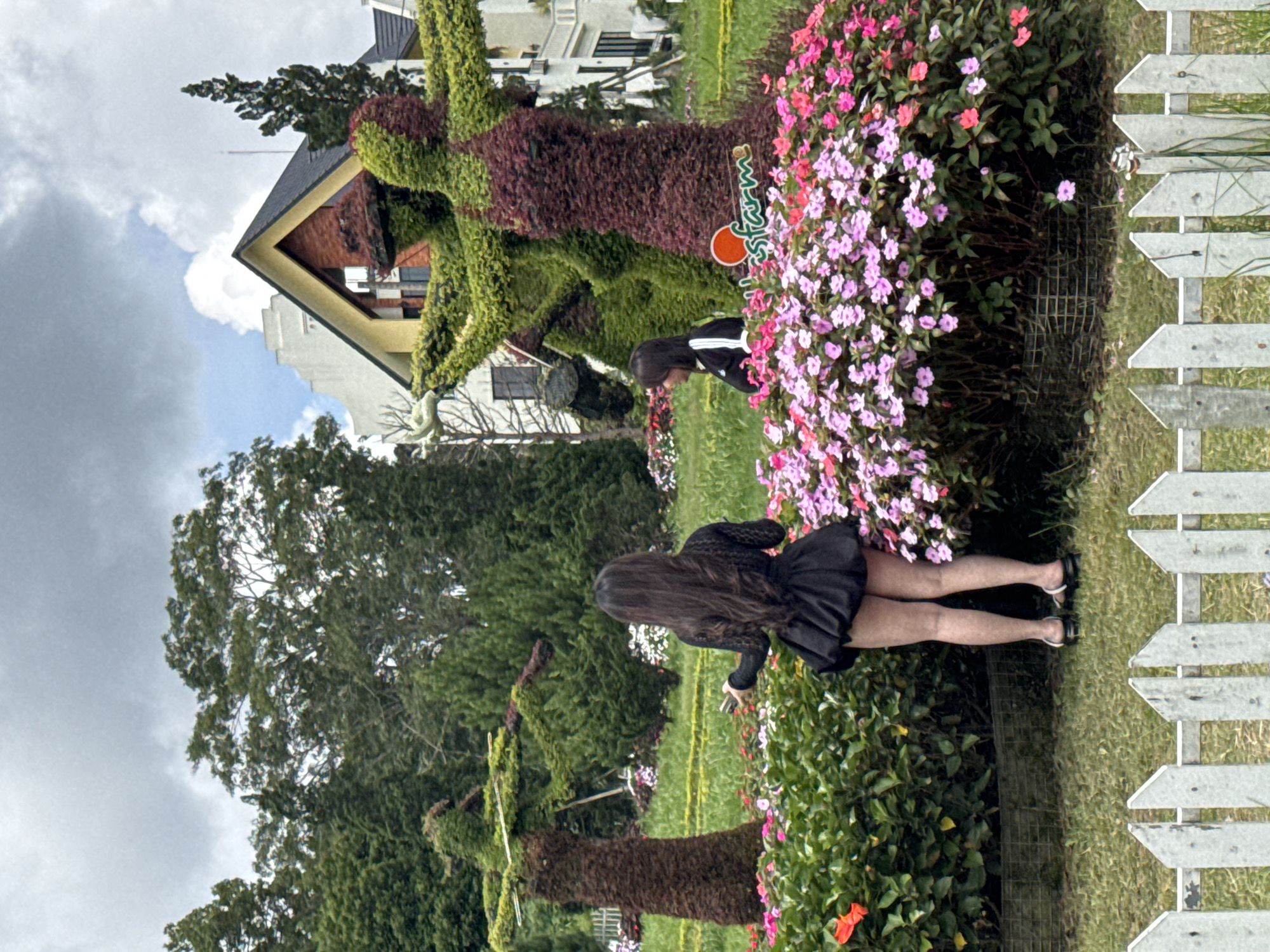 The image depicts a park-like setting with lush greenery and floral arrangements. In the foreground, a white picket fence runs horizontally across the bottom of the frame. Behind the fence, a woman with long dark hair is standing in front of a dense cluster of pink and purple flowers, wearing a short black dress and black sandals. She is holding a black object in her right hand. To her right, another person with shorter dark hair is partially visible, facing left. In the background, there are large topiary sculptures that resemble human figures, one on the left and a larger one on the right, both made of dark brown and green plant material. Behind the topiaries, a white building with a distinctive triangular roof and a smaller white building are visible. Trees and a cloudy sky form the backdrop. The presence of topiary art and the European-style architecture suggest this location is likely within the grounds of a tourist attraction in Da Lat, Vietnam, potentially a flower garden or a themed park such as the "Lost Farm."