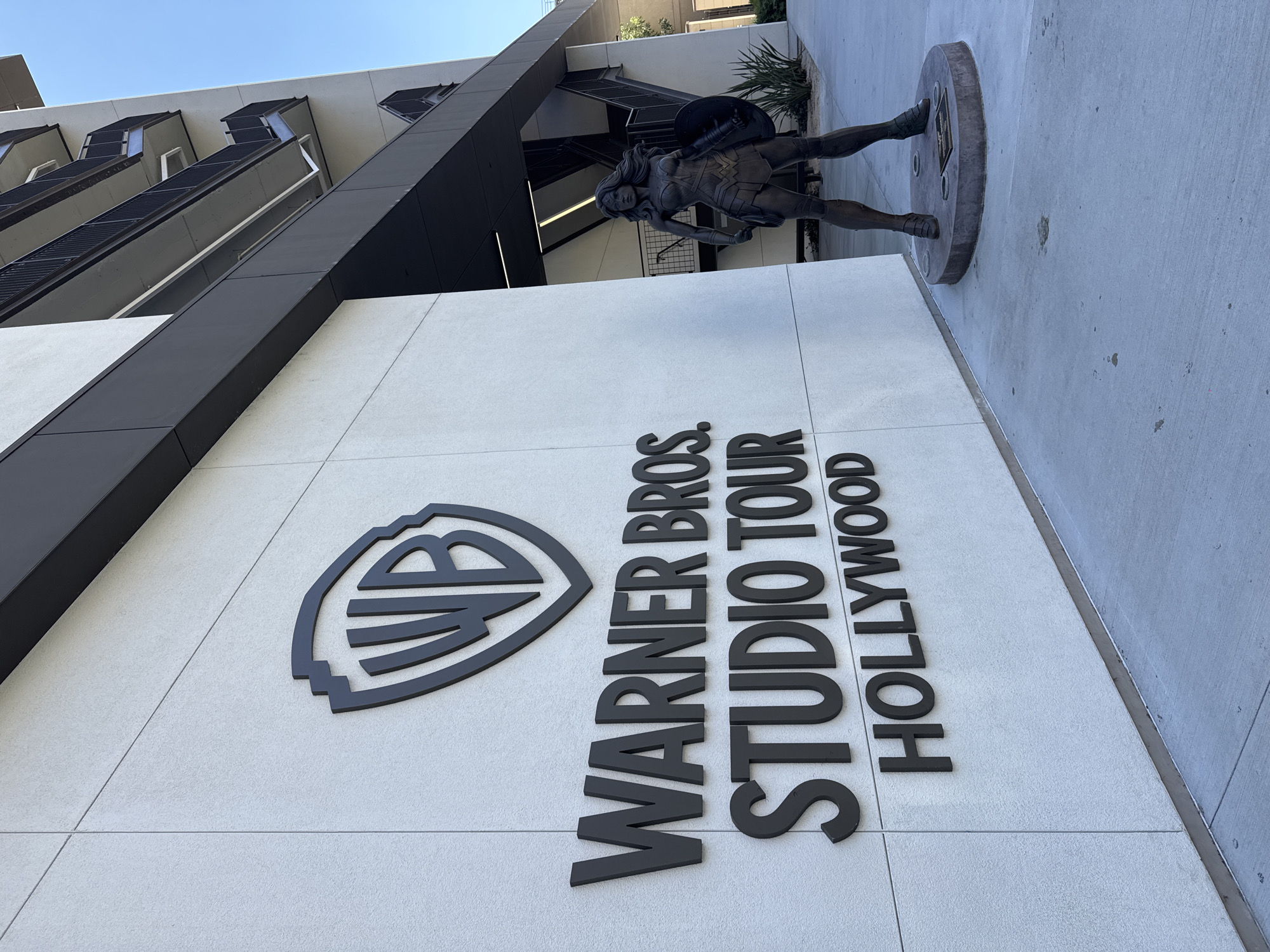 The image depicts the exterior of the Warner Bros. Studio Tour Hollywood in Burbank, California, United States of America. The facade of a modern building features large, dark grey, three-dimensional lettering that reads "WARNER BROS. STUDIO TOUR HOLLYWOOD". Above this text, the iconic WB shield logo is prominently displayed. To the right of the building, a bronze statue of Wonder Woman stands on a circular pedestal. The statue is positioned in a heroic stance, holding a shield and a sword. The ground is paved with concrete, and a clear blue sky is visible in the background. The environment appears to be an outdoor walkway or plaza outside the studio.