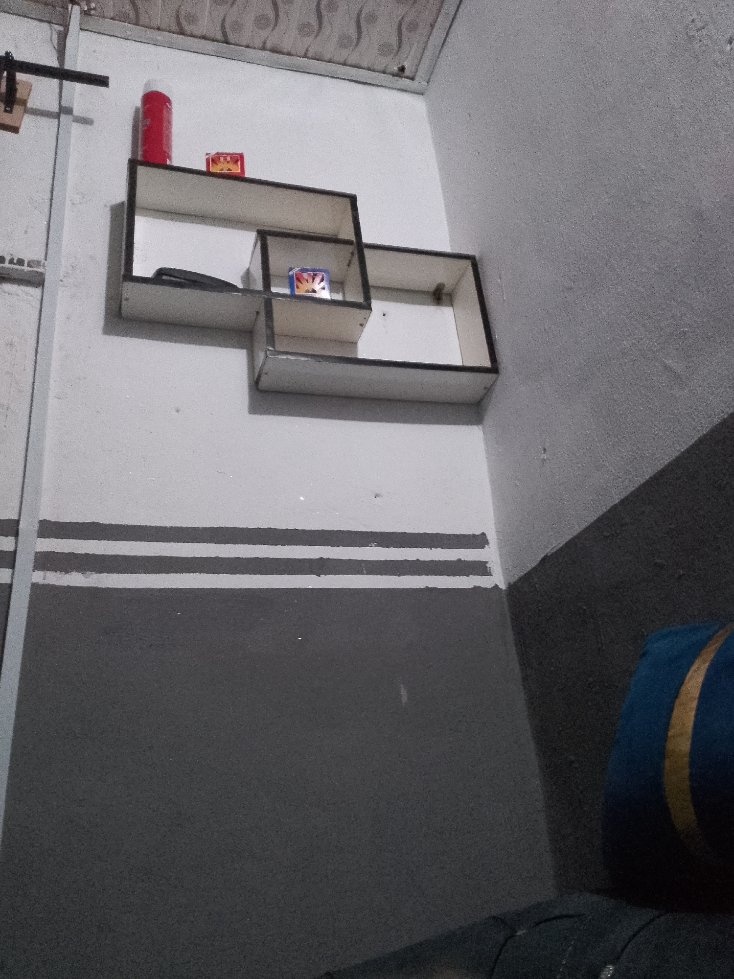 The image shows a shelf mounted on a wall. On the shelf are a red can and two small boxes. Below the shelf are two horizontal gray stripes painted on the wall. The wall is painted white above the stripes and dark gray below. A blue and gold pillow is visible in the lower right corner of the image. The overall scene suggests an indoor domestic setting, potentially in Jauro Yinu, Nigeria, given the prompt's context.