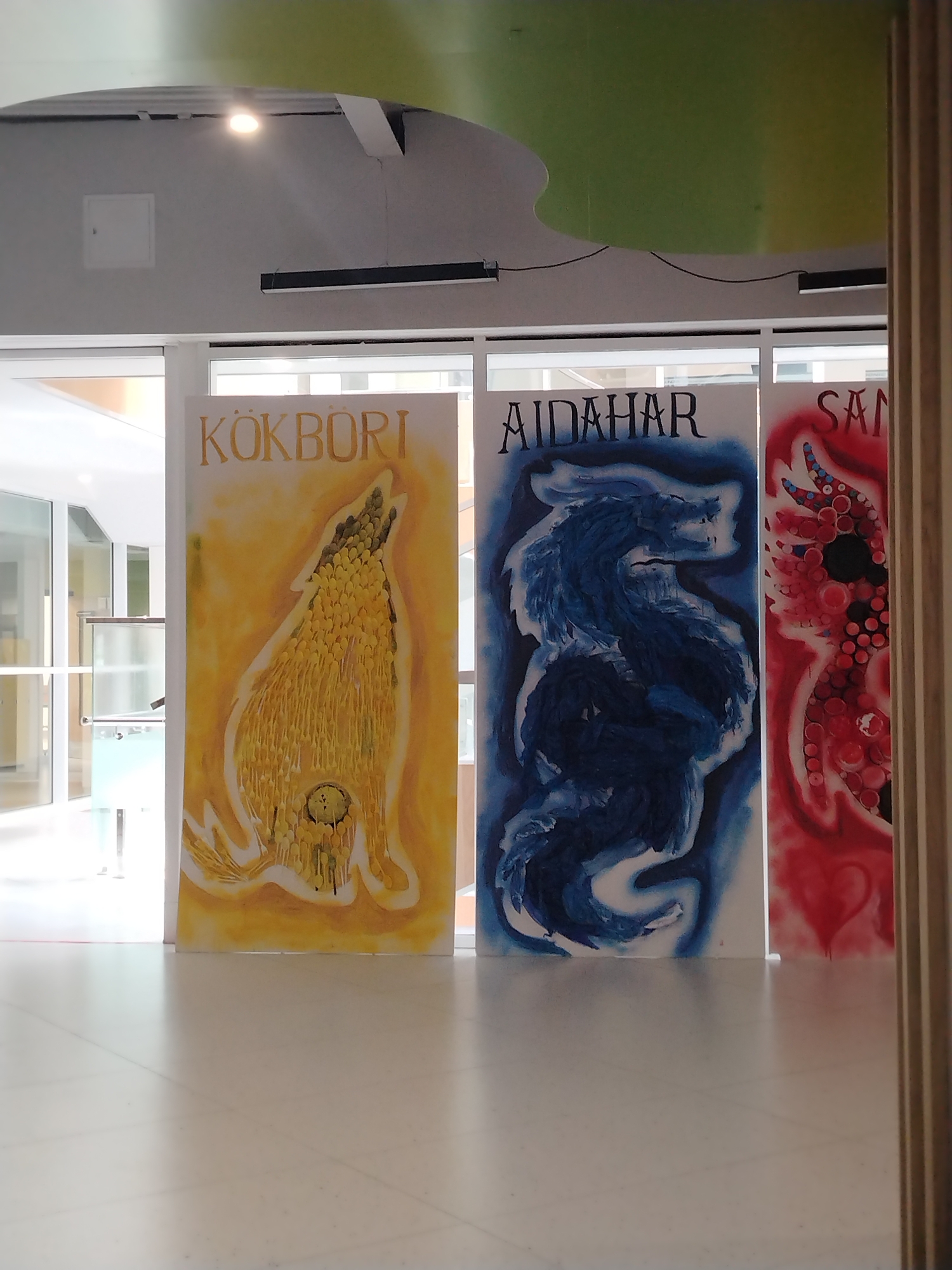 The image shows three large vertical paintings displayed indoors, possibly in a public space or gallery, with a light-colored tiled floor in the foreground and a ceiling with lighting fixtures and a curved green and yellow design overhead. The paintings are:
1. **Left painting:** Features the word "KÖKBÖRI" in a stylized font at the top, followed by a yellow and orange illustration of a creature that resembles a wolf or a mythical canine with its mouth open, howling. The creature is rendered with many small, overlapping yellow elements, giving it a textured appearance.
2. **Middle painting:** Displays the word "AIDAHAR" in a similar stylized font, with a dark blue, almost black, illustration of a dragon or serpent-like creature. The creature is depicted in a dynamic, coiled pose.
3. **Right painting:** Shows the word "SAN" in a stylized font, followed by a vibrant red illustration of a creature with multiple pointed appendages, resembling antlers or fins, decorated with black and red circular patterns. A red heart shape is visible at the bottom of this painting.
The paintings are placed adjacent to each other, and the scene is well-lit. The architectural style of the background, including large windows, suggests a modern building. The presence of a light-colored tiled floor and the overall clean aesthetic implies an indoor environment. There are no people visible in the image.
The location is the National Museum of the Republic of Kazakhstan in Astana, Kazakhstan. The paintings are part of an exhibition, and their thematic content likely relates to Kazakh mythology or folklore, given the names and imagery presented. "Köktöri" could refer to a mythological sky deity or spirit, "Aidahar" is likely a variation of "Ajdaha," a dragon or serpent in Turkic mythology, and "San" is a common word in many languages, but in this context, it could be part of a name or title related to another mythical figure or concept.