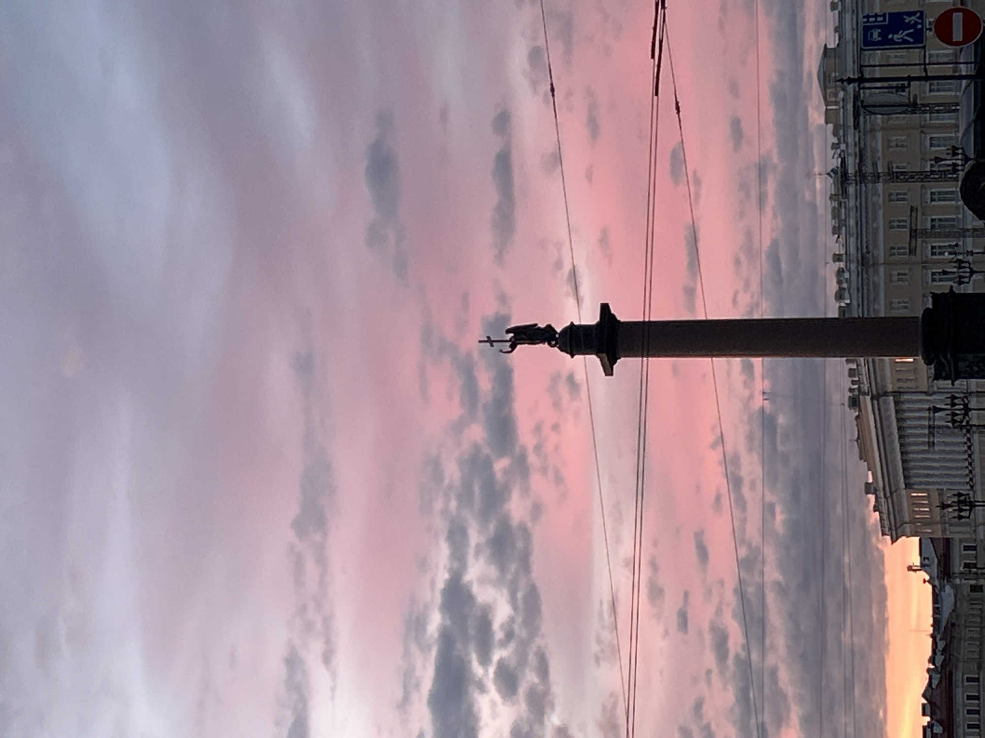 In an urban setting under a soft pink and purple sky at dusk, a tall, slender column topped with a statue of an angel holding a cross stands prominently in the foreground. The column is dark and cylindrical, with the angel silhouette visible against the colorful sky. Below the column, a row of classical buildings with ornate facades, including columns and arched windows, stretches across the mid-ground. Power lines are strung horizontally across the scene, partially obscuring the sky and buildings. In the distance, the faint outlines of other structures are visible. The overall atmosphere is serene and majestic, with the dramatic sky providing a backdrop to the historical architecture and the central monument.