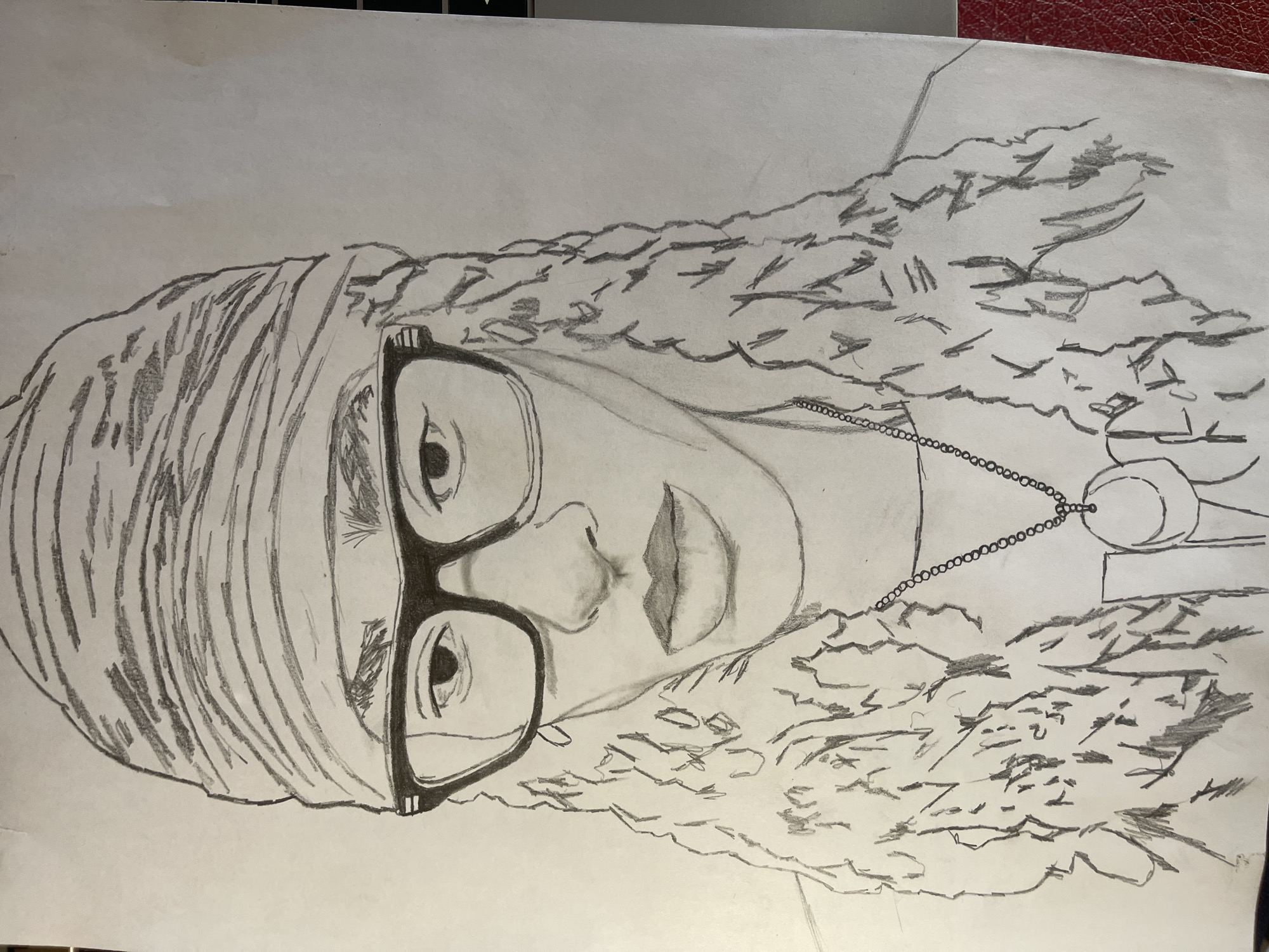The image displays a head-and-shoulders pencil drawing of an individual on a white sheet of paper. The subject, possibly young, is depicted with long, curly or wavy hair flowing down the sides, partially obscuring the shoulders. A textured headwrap or turban covers the top of the head. The individual wears thick-rimmed, dark, rectangular eyeglasses. Facial features include well-defined eyebrows, distinct eyes, a shaded nose, and full lips. A necklace with a circular pendant, which may outline the letters "JO" or a similar symbol, hangs around the neck over a faintly sketched garment. The artwork uses varied line weights and shading to create texture and depth. The background is plain white paper. No specific physical location within the city of Jos, Nigeria, or elsewhere, is discernible from the content of this drawn image. The drawing appears to be photographed on a flat surface, with a dark edge visible on the right and a small section of a reddish, textured surface in the bottom right corner of the frame.