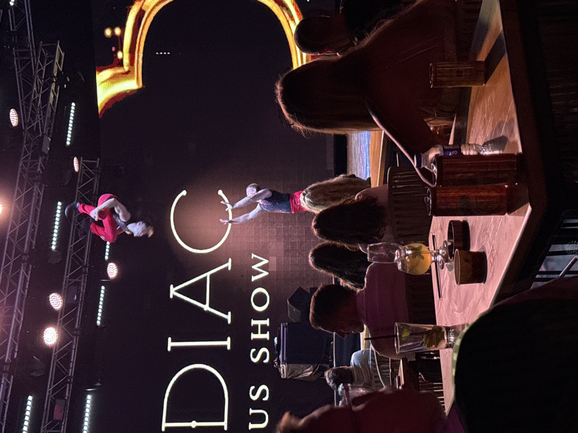 An indoor aerial performance is taking place in Bodrum, Turkey. A male performer in a red costume is suspended in the air, seemingly balanced by his feet. Below him, another male performer in a black top and red skirt is standing on stage, holding his arms up. Behind the performers, the words "DIAC US SHOW" are displayed in large white letters against a dark background. To the right, a stylized golden "C" graphic is visible. In the foreground, several audience members are seated at tables, some with drinks and cans on them. The lighting appears to be stage lighting, with spotlights illuminating the performers.