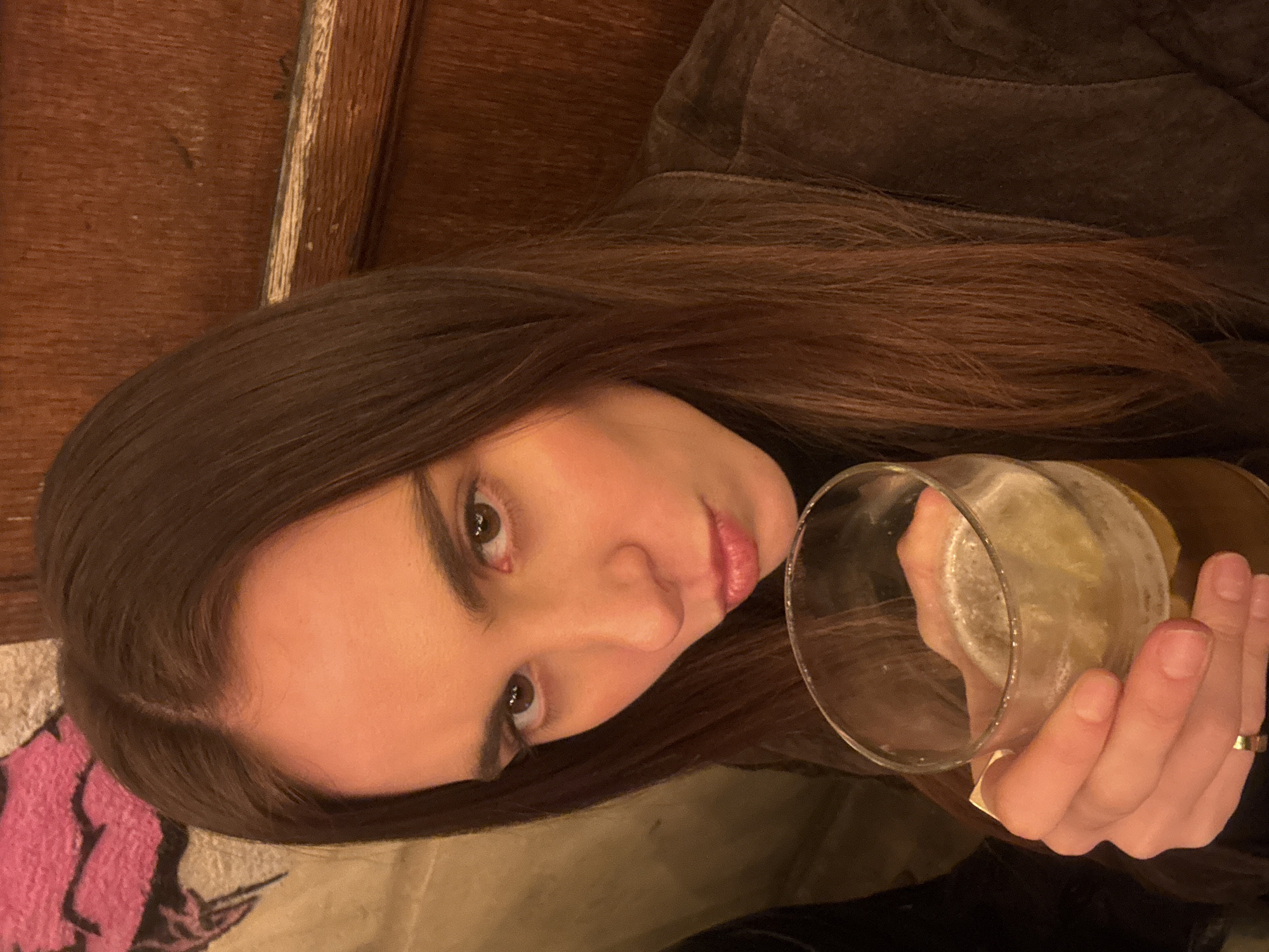 A woman with long brown hair holds a glass of a light-colored beverage. She is wearing a brown jacket. The background includes a wooden surface and a portion of a pink patterned textile. The exact location in Paris, France, cannot be determined from the image.