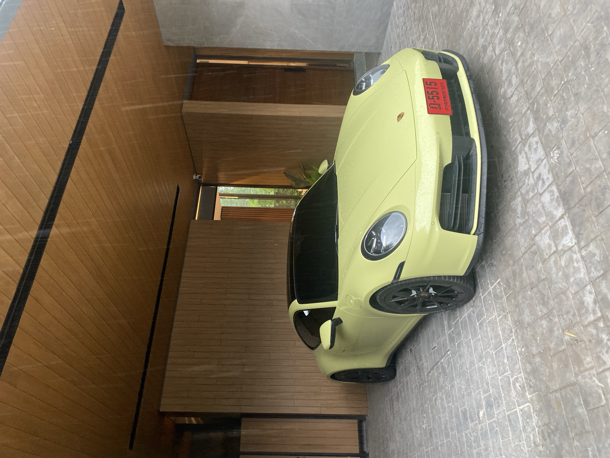 The image shows a pale yellow Porsche parked outside a building. The license plate reads "ข 5515 กรุงเทพมหานคร", indicating that the car is registered in Bangkok, Thailand. The building is constructed from wood, with a grey stone pathway. The location is Choeng Thale, Thailand. No people are visible. It appears to be raining.