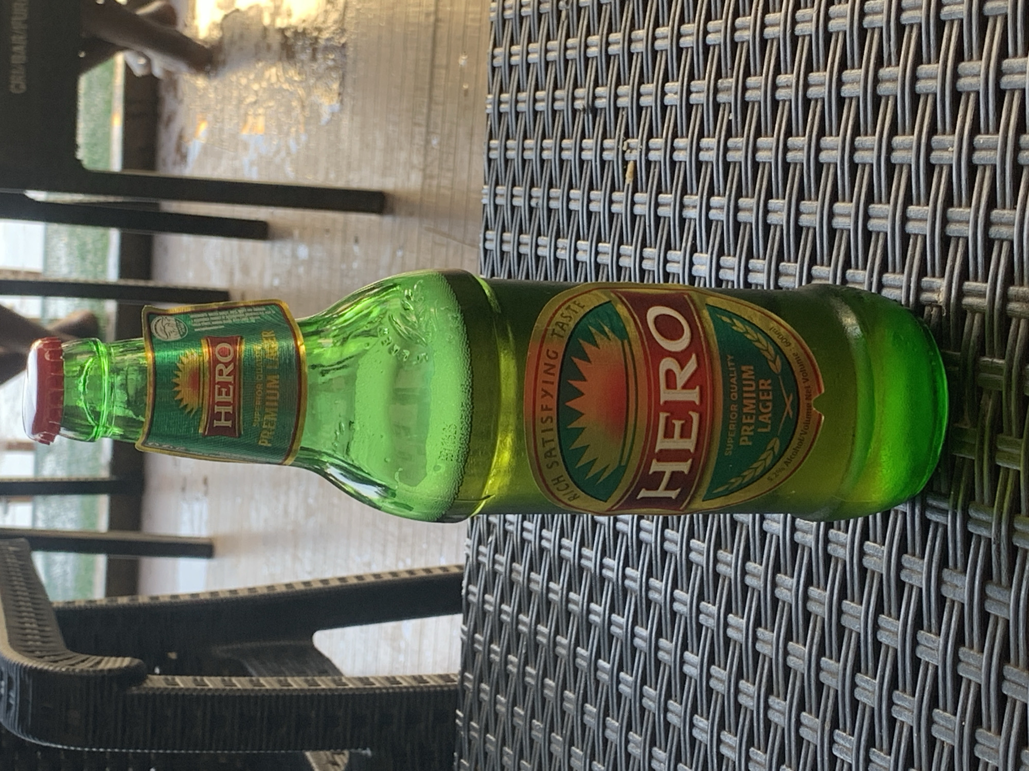 A green glass bottle of "Hero Premium Lager" sits horizontally on a dark gray wicker-patterned surface, likely a table. The bottle is capped with a red metal cap. A black plastic chair is partially visible in the lower left and upper left corner of the frame. The background features a blurry, light area, possibly indicating an outdoor setting. There is no specific information about Barakin Lerawi, Nigeria, visible in the image.