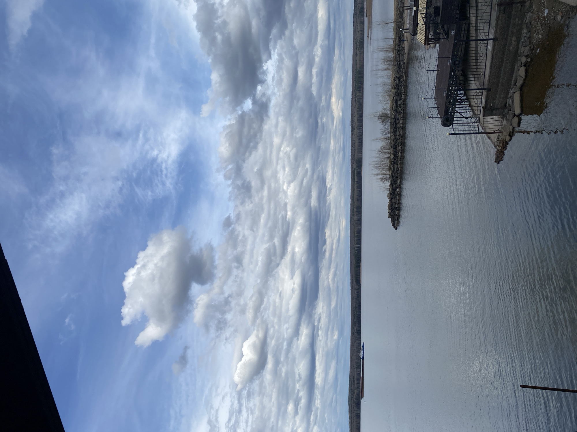 This media file presents a panoramic view of a wide body of water, likely a river or a large lake, under a dynamic sky. The scene is oriented horizontally, depicting a serene waterfront landscape.
The sky occupies a significant portion of the image, showcasing varying cloud formations against a light blue backdrop. There are wispy, high-altitude clouds, as well as more defined, fluffy white cumulus clouds. Towards the center and right, darker, elongated clouds form distinct rows, possibly altocumulus or stratocumulus, indicating a partly cloudy but bright day.
Below the sky, a vast expanse of calm, greyish-blue water stretches across the frame. The water reflects the clouds and sky, and subtle ripples are visible on its surface. A distant shoreline, lined with what appear to be bare trees or sparse vegetation, marks the horizon across the water.
In the mid-ground on the left, a small, dark boat is visible on the water. The foreground on the right features a developed shoreline with a structure that appears to be a pier or a dock. This area is characterized by rocky retaining walls along the water's edge. The pier has dark metal railings with decorative elements, and possibly some lighting fixtures. A dark structure, potentially part of a building or a large gate, is partially visible in the upper right corner of the foreground. On the far left of the foreground, a dark, vertical element, possibly a wooden or metal post, juts out into the water.
The lighting suggests it is daytime, possibly late morning or early afternoon. The bare trees and the overall subdued color palette hint at a cooler season, such as late winter or early spring. No people or visible text are present in the image. The scene conveys a sense of peaceful solitude at a riverside or lakeside location, consistent with views found along the Kama River in regions like Chaikovsky, Russia.