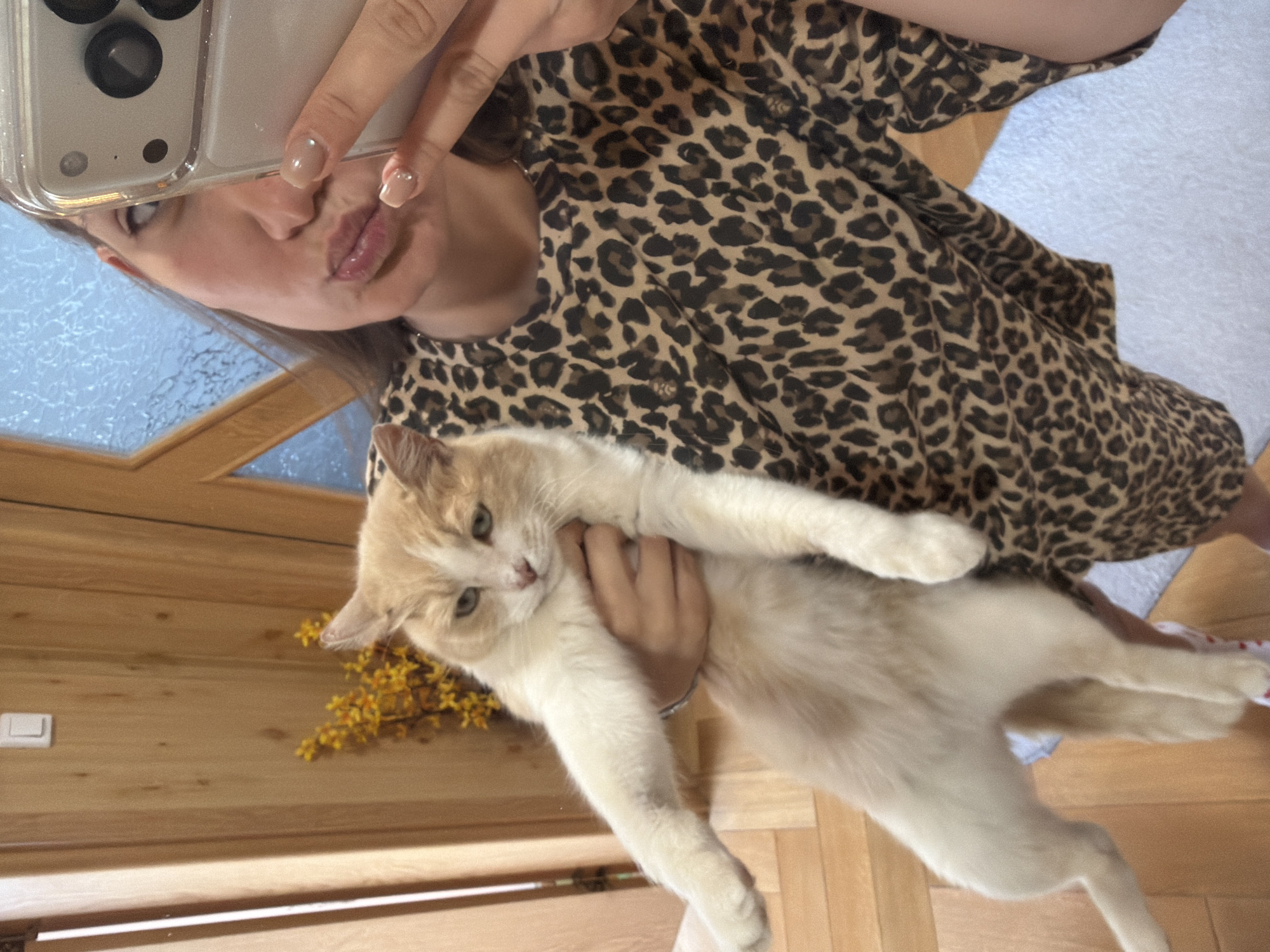 A woman takes a selfie with her cat in what appears to be an indoor setting, possibly in a home. The woman, wearing a leopard print top, holds a cream-colored cat with some orange patches. The cat is facing the left and looking slightly downward. The background includes a wooden frame, possibly a window or door, and some yellow flowers are visible to the left of the cat. A portion of the woman's face and her hands holding a phone are visible in the upper left of the frame. The scene appears to be in Buhovo, Bulgaria.