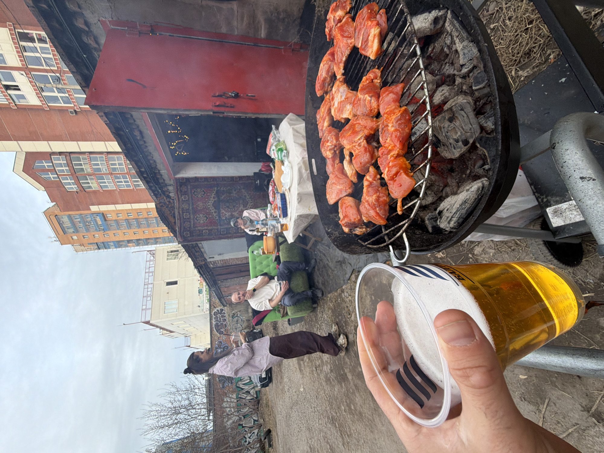 The image captures an outdoor scene in what appears to be Krasnoyarsk, Russia. In the foreground, a hand holds a plastic cup filled with beer. A barbecue grill with skewered meat sits to the left. In the background, there are two men in casual clothing are seated on a green couch, situated under what looks like a rug hanging from the edge of a building. The buildings in the background are brick. In the mid-ground is a woman standing near the green couch and barbecue grill. The scene is set outdoors, with visible graffiti and what appears to be a fence in the distance.
