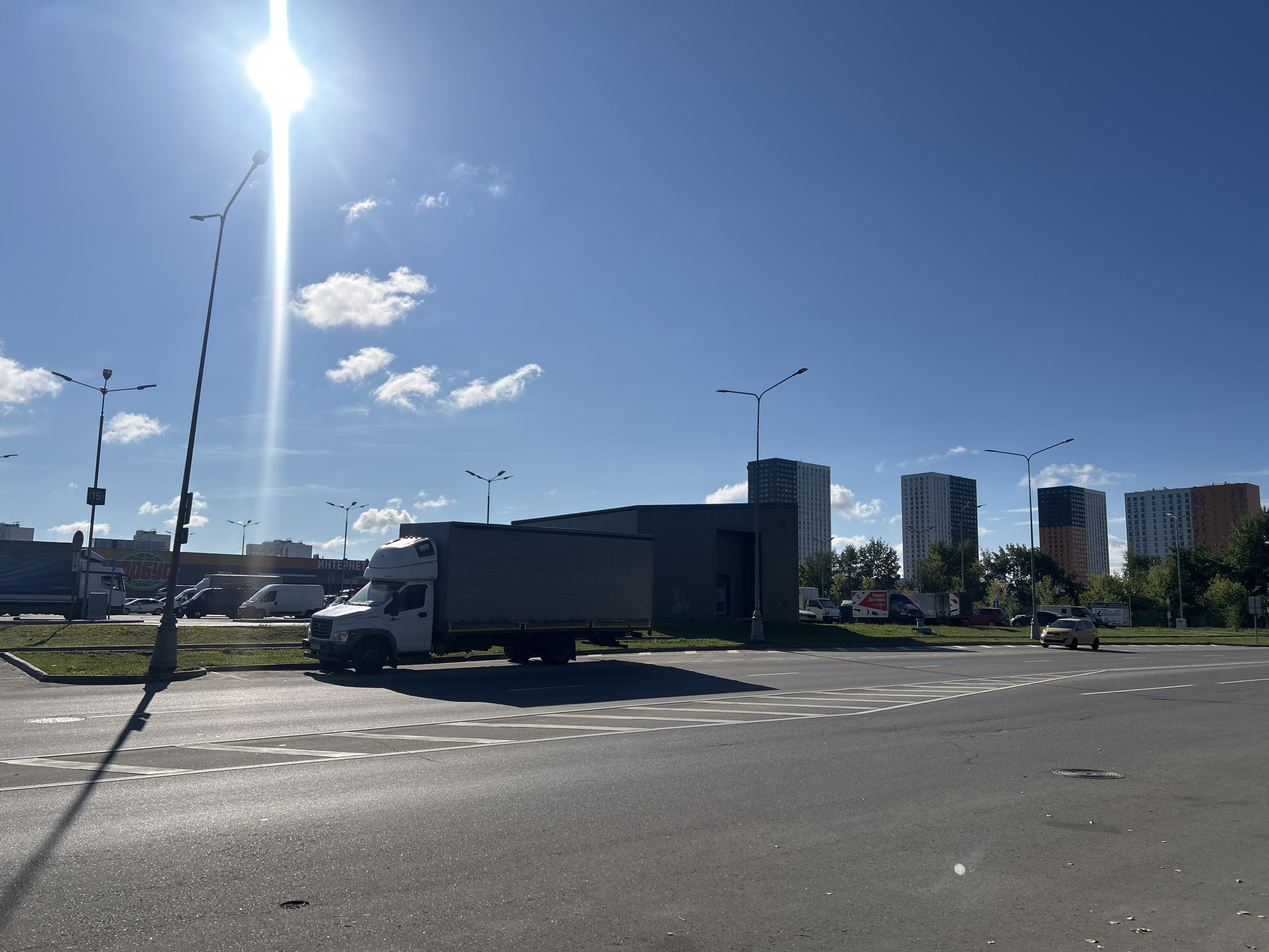 The image depicts an urban street scene on a sunny day with a clear blue sky and scattered white clouds. In the foreground, a multi-lane asphalt road with white markings is visible. A street lamp casts a long shadow across the road.
In the middle ground, a white truck with a grey cargo box is parked on the side of the road. To the left of the truck, a dark-colored building is situated, with a grass area in front. Several other vehicles, including vans and cars, are parked in the background. Some commercial buildings and signs, one with Russian text, are visible.
In the background, a row of tall, modern apartment buildings, mostly in shades of grey, white, and orange, dominate the skyline. Trees and more buildings are scattered throughout the distance.
The scene appears to be a commercial or mixed-use area within a city, potentially on the outskirts of Moscow, Russia, given the architectural style of the buildings and the presence of Russian text on a sign. There are no people clearly visible in the image. The overall impression is one of a typical urban environment on a bright, clear day.