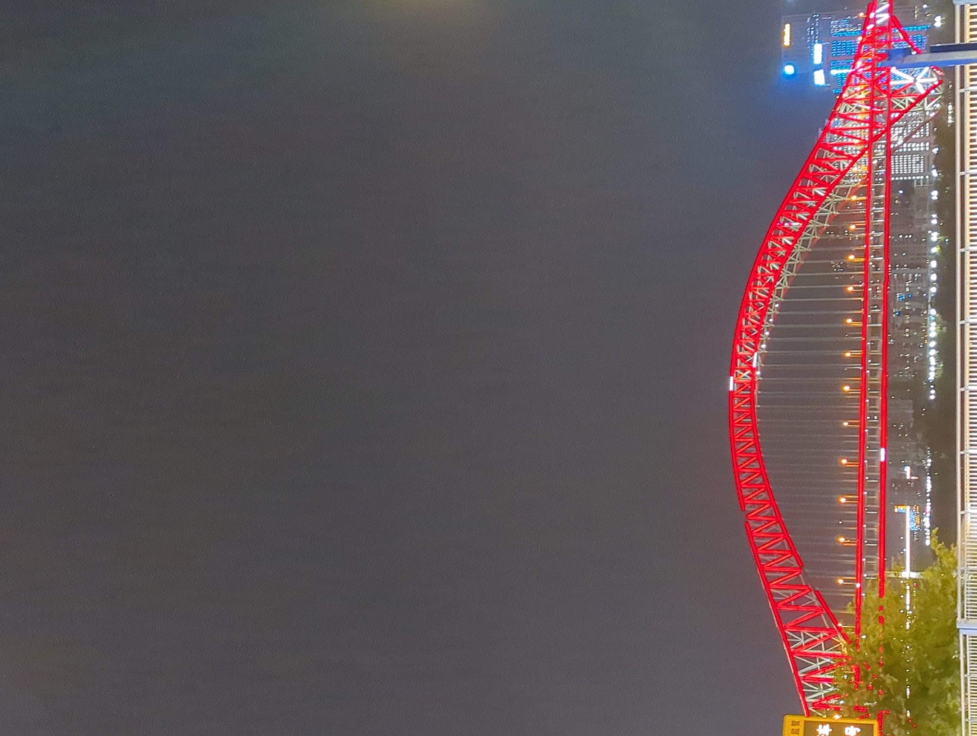 A red arched bridge is illuminated against a dark sky. The bridge has a red lattice structure and is supported by numerous vertical cables. Lights are visible along the length of the bridge and also in the background, suggesting a city environment. The lower part of the image shows some greenery in the foreground. The scene appears to be a nighttime cityscape, likely in the vicinity of Jiangbeicheng Street in China, given the context.