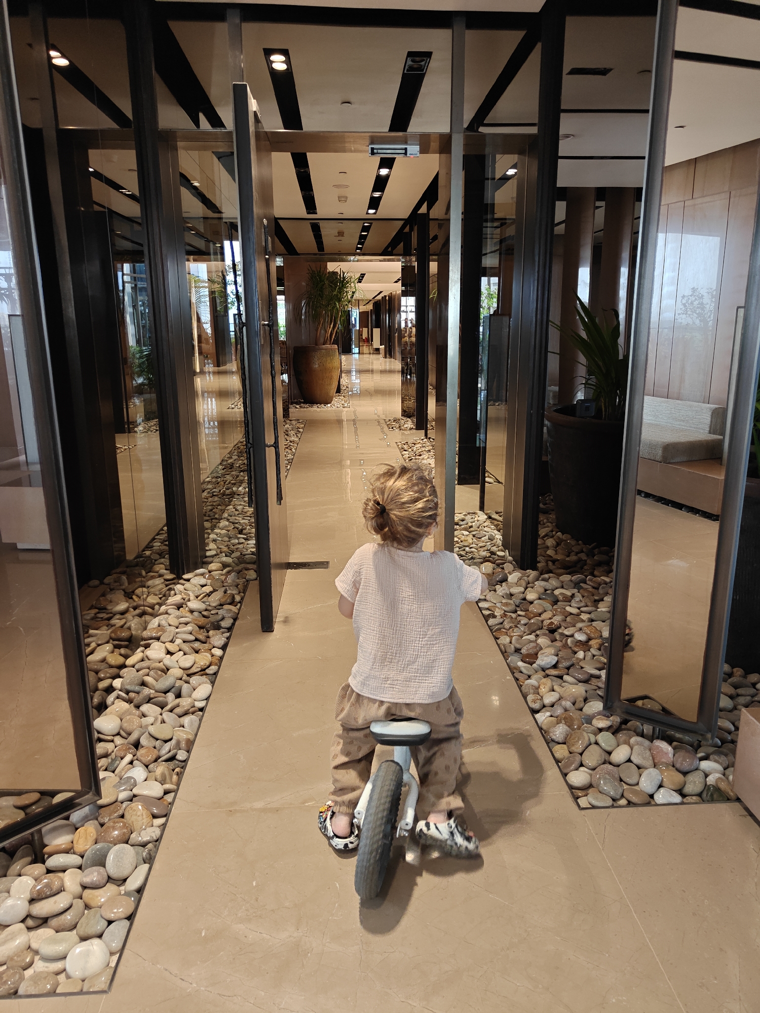 A child rides a balance bike down a corridor in what appears to be a modern interior space. The child is facing away from the camera, wearing a light-colored shirt and pants, and their hair is gathered in a ponytail. The corridor is lined with floor-to-ceiling panels that seem to be either mirrors or glass, flanked by decorative pebbles along the edges. There are potted plants in the background and the corridor leads to an open space further ahead.
The corridor's architectural style, interior design, and decorative elements, combined with a security sign on the ceiling, suggest the scene could be a luxurious lobby in Mumbai, India.