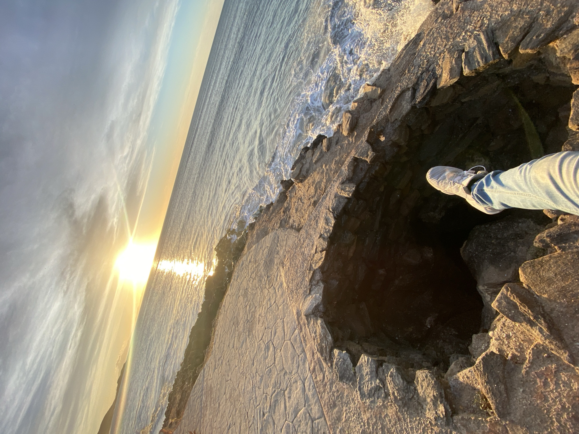 A medium shot depicts a stone pier abutting the ocean, with a round hole cut into the pier's surface. The sun shines brightly on the water and pier, creating strong shadows. Part of a person's leg and foot, clad in blue jeans and a white sneaker, is visible at the edge of the hole. The image is tilted to the left. The location is likely Juan Griego, Venezuela, given the prompt.
