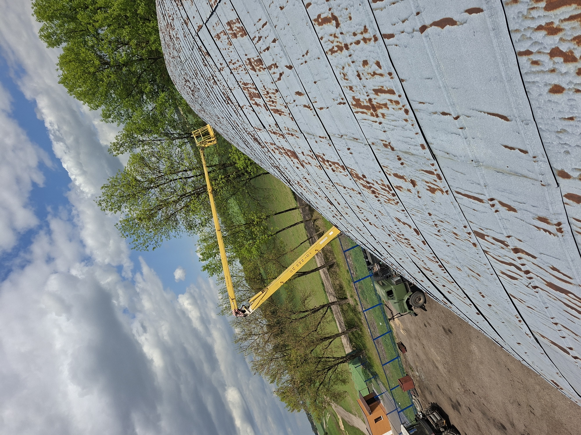 The image shows a large, curved metal structure, possibly a grain silo, with visible rust. A yellow articulated boom lift is positioned next to it, its basket reaching toward the top. Trees with green foliage are visible in the background and partially obscuring the structure. The sky is partially cloudy with blue patches. An army vehicle is visible near the structure. The location is Zhabinka, Belarus.