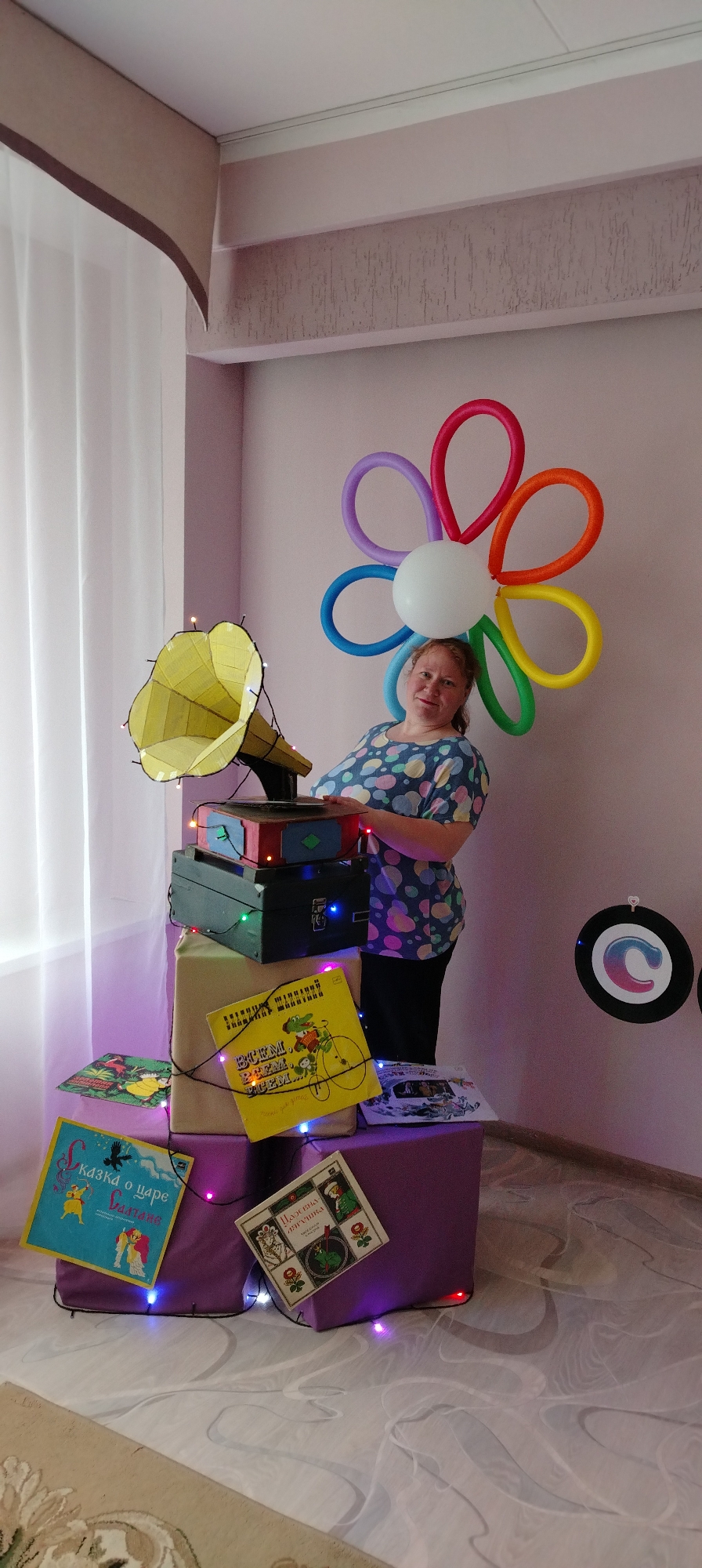 The image shows a woman standing next to a stack of decorated boxes and books in a room with light pink walls. The room has a window with white curtains. The location is likely Chaikovsky, Russia, given the Russian book covers visible.
* **People:** A woman with fair skin and reddish hair is standing to the right of the stack. She is wearing a blue and purple patterned shirt and dark pants. She is smiling and has her hand resting on top of the stack.
* **Subjects:** The main subjects are the woman and the decorated stack, which includes a homemade gramophone with a yellow horn, various wrapped boxes, and Russian children's books.
* **Objects:** Key objects include:
* A homemade gramophone with a yellow horn.
* Wrapped boxes, stacked on top of each other.
* Russian children's books with titles in Russian on the covers ("Сказка о царе Салтане", "Царевна лягушка", "ВРЕМ, ВРЕМ, ВРЕМ...")
* String lights wrapped around the stack.
* A balloon flower decoration behind the woman.
* Decorations on the wall in the upper right corner of the image.
* **Actions:** The woman is posing for the photo, with her hand placed on the stack. The stack is arranged to create a visually appealing display.
* **Interactions:** The woman interacts with the stack by resting her hand on it.
* **Environment:** The scene is set indoors in a room with light pink walls. There is a window with white curtains on the left side of the image. The floor is a light marble or tile pattern. There appears to be a carpet partially visible in the bottom of the image. The lighting is bright, and the overall mood is festive.