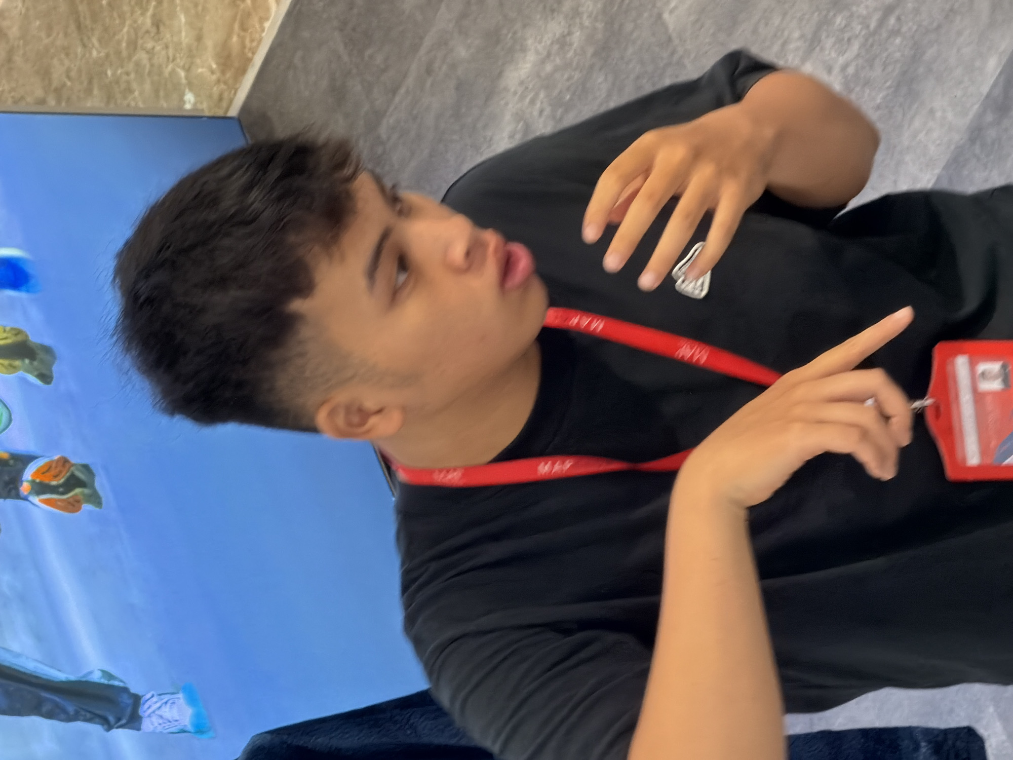 A young person with dark, short hair is captured from the chest up, looking to their right and speaking. They are wearing a black t-shirt with a red lanyard around their neck, a white logo on the shirt, and a red badge clipped to the lanyard. Their left arm is bent and resting on their chest, while their right hand is raised, with fingers extended, as if emphasizing a point. The background shows a wall with a marble-like texture and a bright blue backdrop with images of feet wearing colorful shoes. The scene appears to be indoors, possibly at an event or exhibition. There is no discernible information in the image to suggest it is located in Nha Trang, Vietnam.
