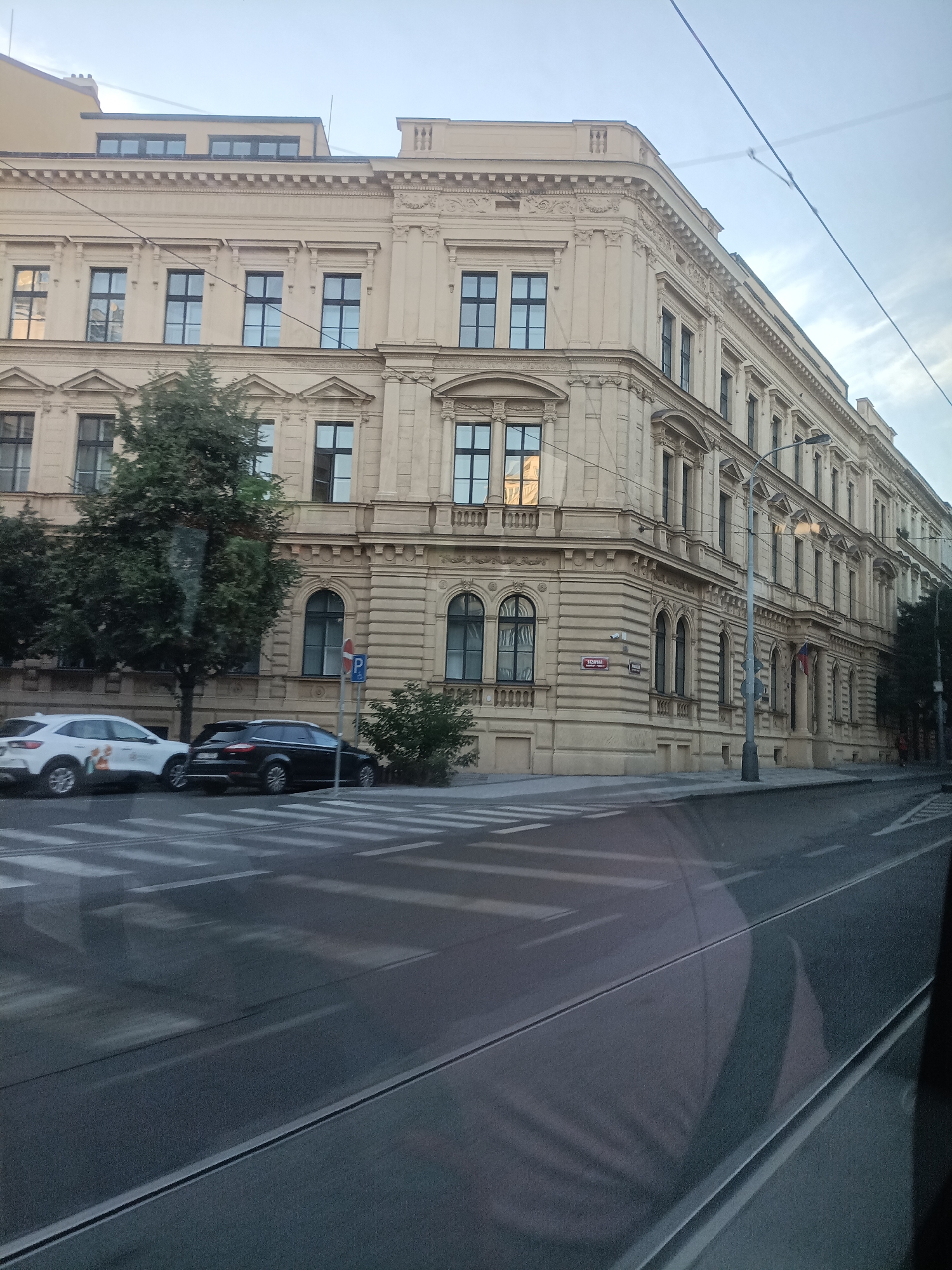 The image shows a street scene in Prague, Czechia, with a grand, light-colored building dominating the right side of the frame. The building has a classical architectural style, featuring multiple stories, numerous windows with dark frames, and decorative cornices. The street in the foreground is paved, with visible white pedestrian crossings. Two cars are parked on the side of the street, a white SUV and a dark sedan. Trees line the sidewalk in front of the building. Overhead, power lines are visible against a cloudy sky. The overall impression is of a well-maintained urban environment with significant historical architecture.
