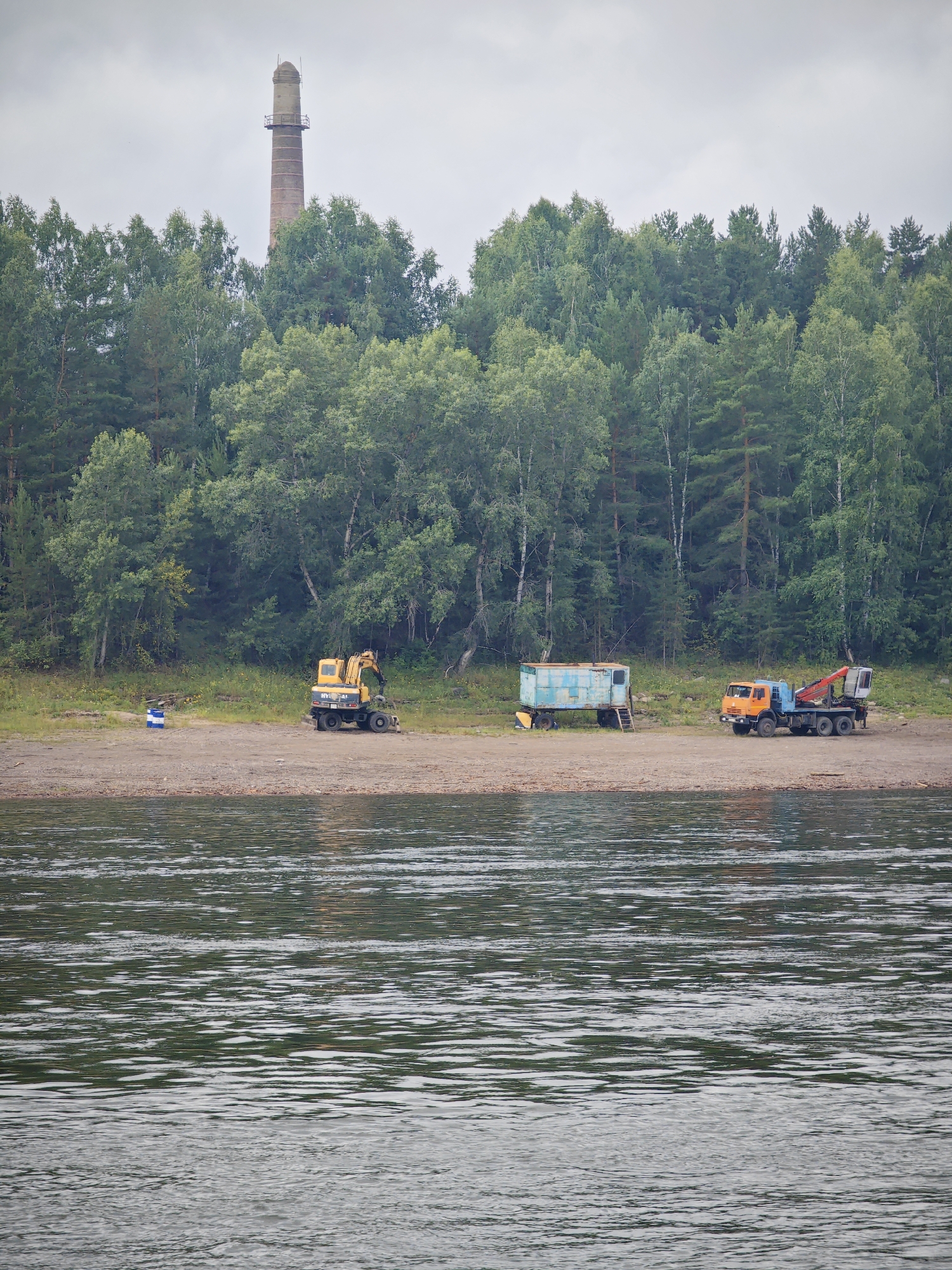 In Lesosibirsk, Russia, on a riverbank, a scene unfolds with a backdrop of dense forest. The foreground features the shimmering surface of a body of water, with ripples creating a textured pattern. On the sandy shore, several pieces of heavy machinery are present: a yellow excavator, a light blue and white trailer, and an orange and blue truck with a crane. In the distance, a tall, red brick smokestack pierces the overcast sky, a solitary industrial remnant against the natural landscape. The overall impression is one of a working waterfront, possibly involved in construction or industrial activity, juxtaposed with the serene wilderness of the surrounding trees.