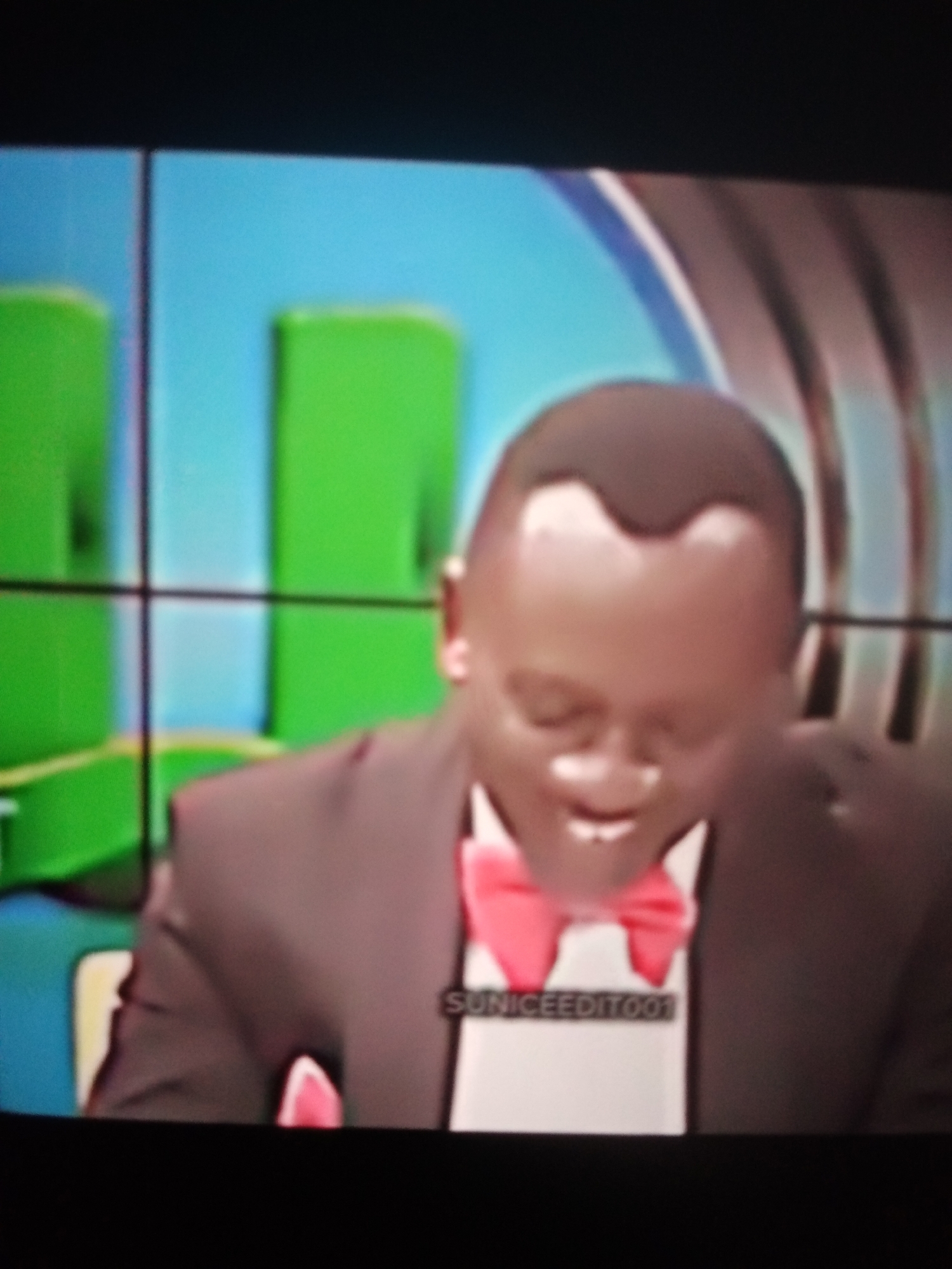The image shows a man wearing a suit and a pink bow tie. He has a broad smile and is looking slightly downwards. The background appears to be a television screen with a blue and green graphic. A watermark with the text "UNICEEDITO01" is visible on the man's shirt. The scene is likely from a television broadcast. There is no identifiable location within the city of Jalingo, Nigeria.