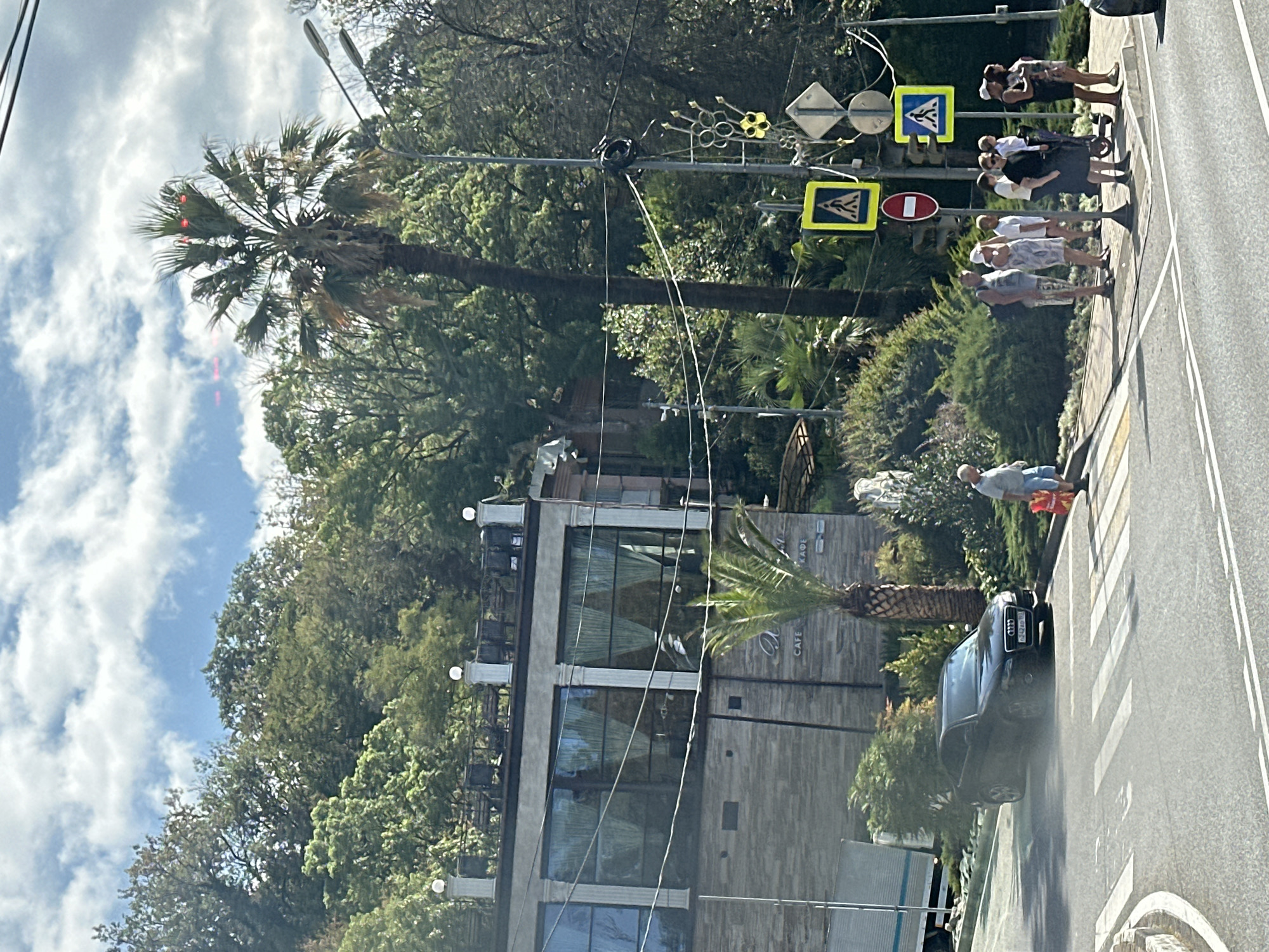 This image shows a street scene in Sochi, Russia, with a modern building featuring large windows and a cafe sign that reads "CAFE". A black Audi car is parked near a crosswalk, and several people are standing on the sidewalk, appearing to wait. Traffic signals and pedestrian crossing signs are visible, along with palm trees and lush greenery, suggesting a warm climate. The sky is partly cloudy.