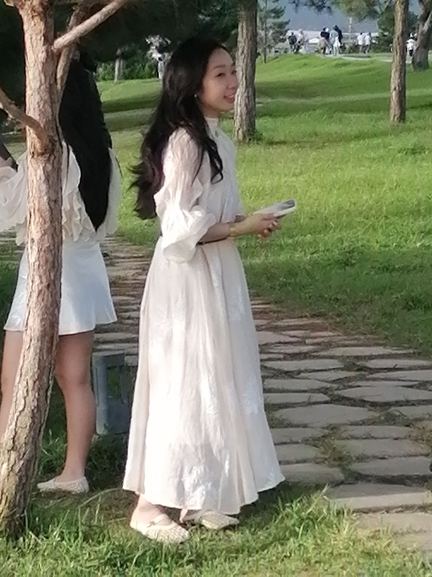 In Da Lat, Vietnam, two women are visible in a park with lush green grass and a paved walkway. A tree trunk is prominently positioned in the foreground on the left. The woman on the right, facing away from the viewer and looking towards the right, wears a long, flowing white dress with bell sleeves and is holding a mobile phone. Her dark hair is long and wavy. To her left, another woman, partially obscured by the tree, is also visible wearing a white dress and appears to be looking forward. The background reveals more trees and a group of people in the distance, suggesting a public outdoor space. The lighting indicates it is daytime, likely late afternoon given the soft sunlight.