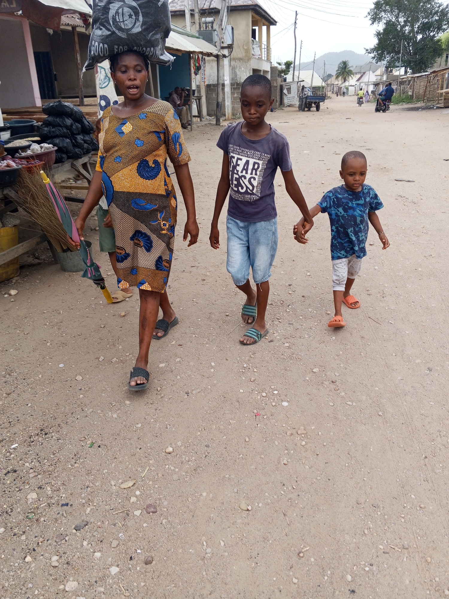 In Adankolo, Nigeria, an adult female walks on a dirt road, carrying a black bag on her head. She wears a patterned dress and dark sandals. Two young boys, one older and one younger, walk beside her, holding hands. The older boy wears a "FEAR LESS" t-shirt and denim shorts, while the younger wears a blue patterned t-shirt and light-colored shorts. The road is lined with buildings and sparse vegetation. Vehicles and pedestrians are visible further down the road.