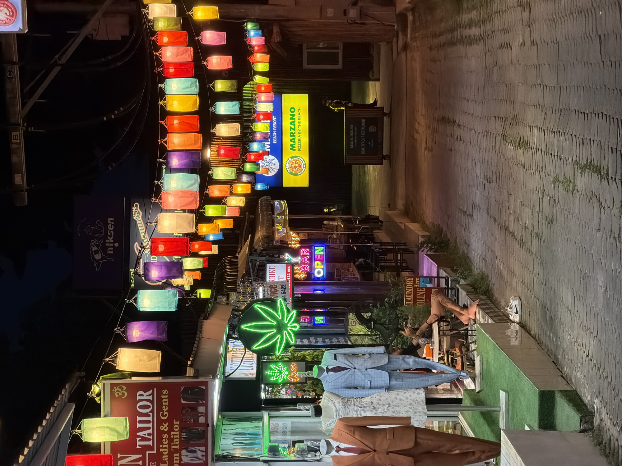 This is a nighttime shot of a narrow street in Baan Chaweng, Thailand, lined with shops and decorated with numerous colorful paper lanterns hanging overhead. The scene depicts a vibrant commercial area with a variety of businesses.
Prominently featured are several signs. One sign reads "MARZANO PIZZERIA BY THE BEACH," another says "MALIBU RESORT BEACH FRONT RESTAURANT," and a "LAUNDRY" sign is also visible. A neon sign with a cannabis leaf design is lit up, and another neon sign advertises "BAR OPEN." There's also a sign for "N TAILOR Ladies & Gents."
Mannequins dressed in suits are displayed outside a shop, showcasing menswear. In the background, people are visible, suggesting ongoing activity in the area. The street is paved with cobblestones. The overall atmosphere is lively and colorful, illuminated by the glow of lanterns and neon signs, indicating it's likely evening or night. The presence of a fan suggests a warm climate, typical of Thailand.