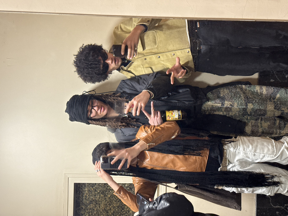 Three individuals are captured in a mirror selfie taken indoors within the city of Paris, France. The scene depicts a casual social interaction, possibly in a hallway or private residence, against a plain, light-colored wall with a recessed panel featuring a textured, woven pattern on the left.
From left to right:
1. A person with dark hair, partially obscured by a black fringed scarf, is taking the photograph. They wear a brown leather jacket, a black top, and light-colored cargo-style pants with a studded belt. Their right hand, adorned with rings, holds a dark-cased smartphone that covers their face. A dark bag is visible on their left side.
2. A person with long, dark dreadlocks or braids is positioned centrally. They wear a black headwrap, eyeglasses, a dark hooded sweatshirt, and a dark puffer jacket, paired with textured camouflage-patterned pants. Their right hand holds a clear glass containing a light-colored beverage, while their left hand displays a "peace" or "V" sign. A yellow-labeled bottle, partially visible, is held beneath the glass by their fingers. A silver bracelet is on their left wrist.
3. A person with dark, voluminous curly hair (afro style) stands on the right, wearing dark sunglasses. They are dressed in a light tan or yellowish suede-like jacket with a collar and chest pockets, and dark-colored pants secured by a prominent studded belt. Their right hand holds a dark-cased smartphone, while their left hand is extended forward in an open, gesturing pose. A ring is visible on their right hand.
All individuals are looking towards the camera, suggesting an intentional pose for the mirror photograph. The lighting is uniform and artificial, characteristic of an indoor setting.