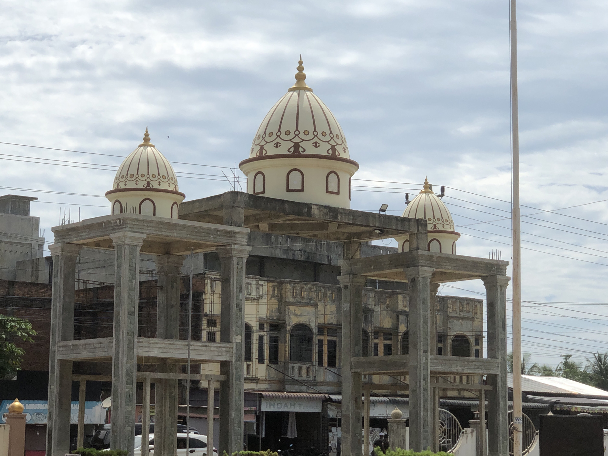 The scene depicts a building featuring multiple domes in Meudangara, Indonesia, partially framed by a concrete structural skeleton in the foreground. Three prominent domes are visible, characterized by their light cream color, reddish-brown intricate decorative patterns, and golden finials. Below each dome, a band of arched, recessed window-like elements is present. The building behind these domes appears older, with a weathered, light yellow facade exhibiting arched windows, balconies, and decorative architectural details. Exposed concrete and rebar suggest ongoing construction or renovation activities on the upper sections of this structure.
In the immediate foreground, an unpainted concrete framework composed of large square columns and horizontal beams forms an open, multi-level structure, partially obscuring the main building. These columns feature simplified capital and base elements. Above the structures, a cloudy, light blue-grey sky is intersected by multiple black power lines.
At ground level, various elements are discernible: parked vehicles, including white cars and motorcycles, are visible. A business sign displaying "INDAH" is present on a lower building section. Small patches of green foliage are scattered throughout the lower portion of the frame. On the far right, a tall, slender white pole extends vertically from the ground, reaching into the sky. A metal gate and a few indistinct human figures are visible in the lower right section of the image.