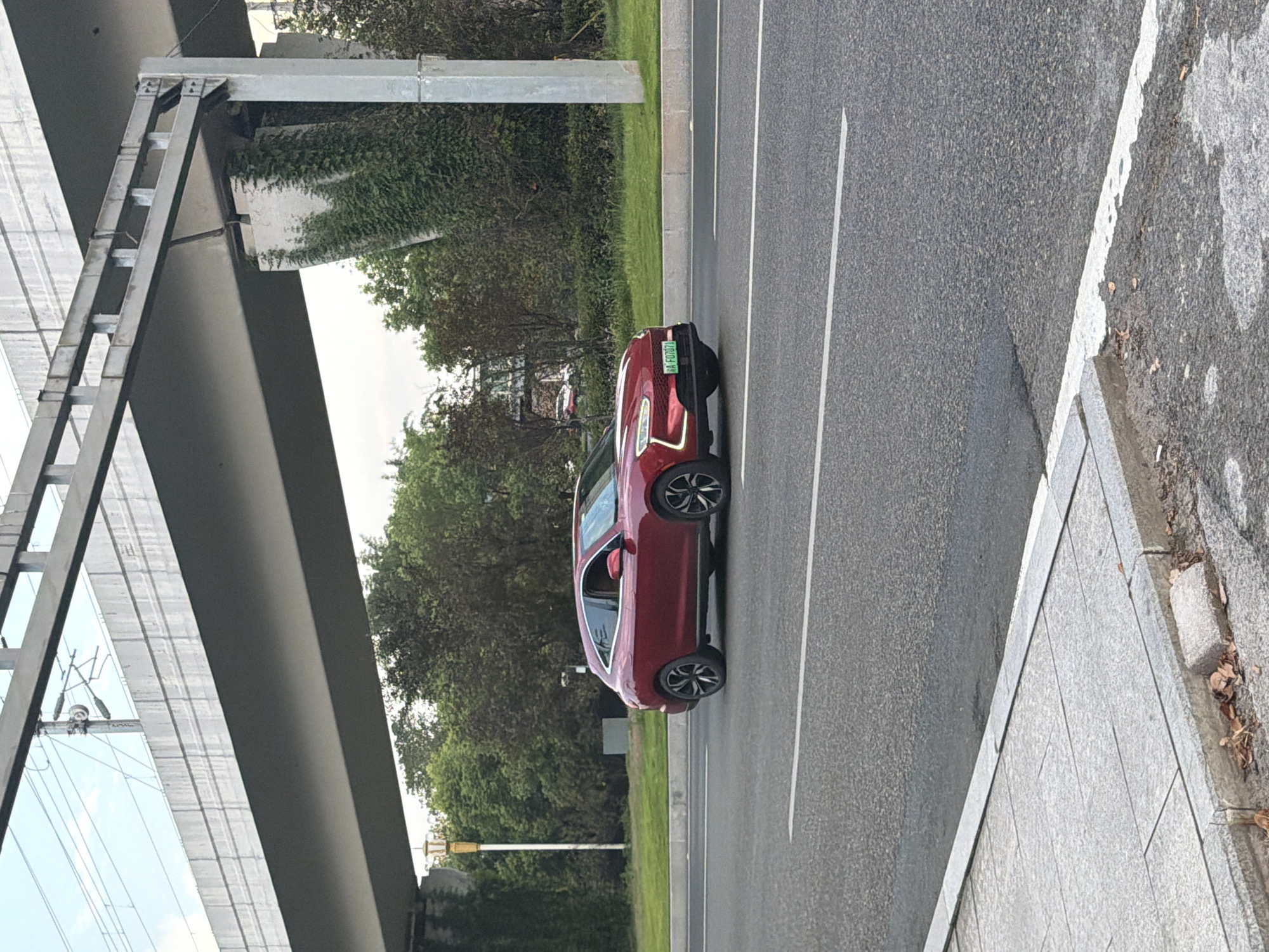 In Jiangjin, China, a red electric vehicle is captured mid-motion on an asphalt road, its license plate reading "A F07071". The car is positioned in the right lane of a multi-lane road. In the background, a grey concrete overpass looms overhead, supported by substantial pillars. Dense green trees and ivy-covered structures frame the scene, suggesting a suburban or semi-urban environment. The sky above is a muted blue with visible power lines crisscrossing the frame, indicating the presence of electrical infrastructure. The foreground features a paved sidewalk constructed of grey stone tiles, separated from the road by a concrete curb. Scattered dry leaves rest along the curb, hinting at the time of year. The lighting suggests it is daytime, likely late afternoon given the soft light.