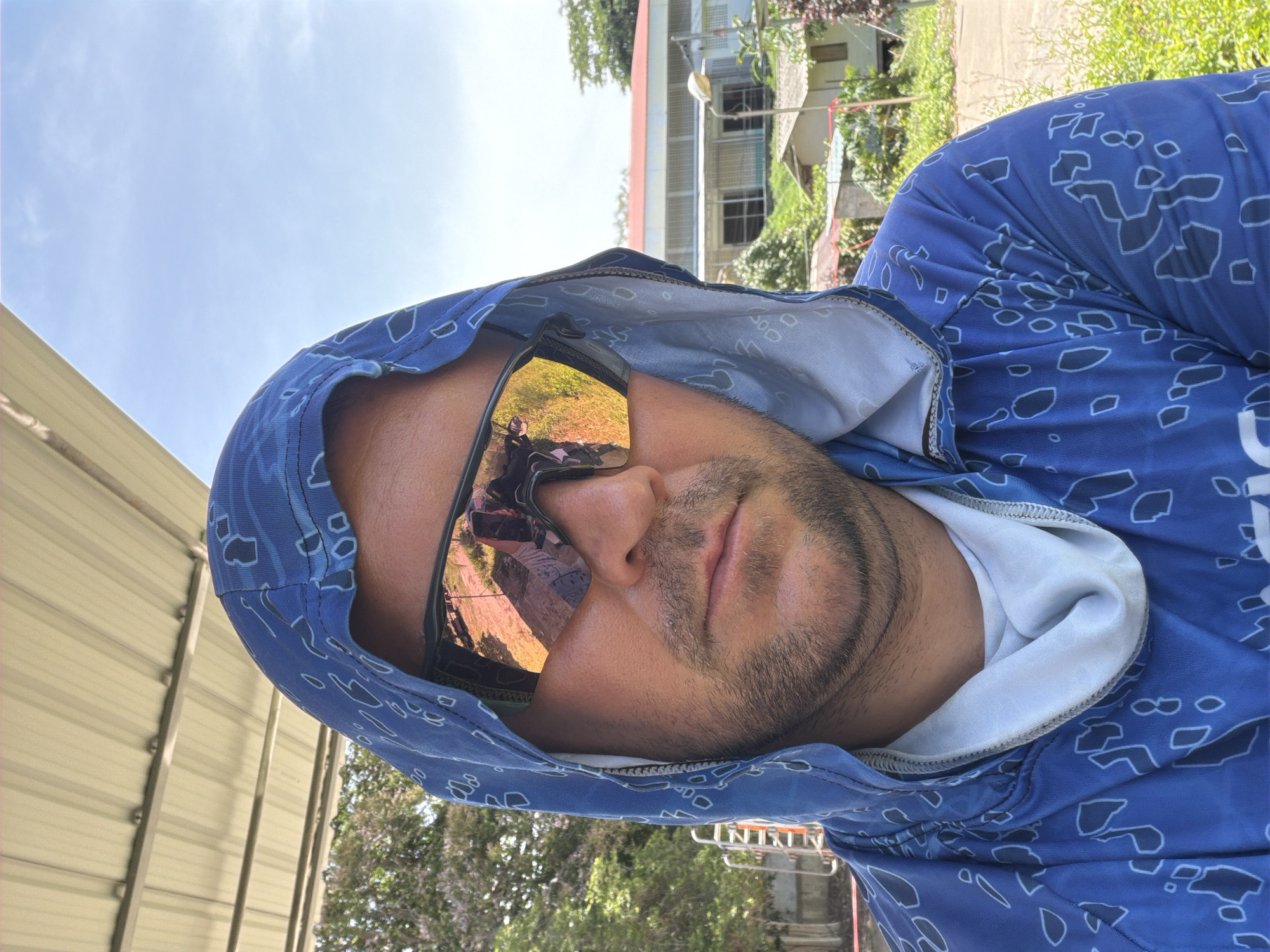 A man wearing a blue hooded shirt with a white insert and sunglasses is taking a selfie outdoors. The sunglasses have a reflective orange tint, showing a reflection of what appears to be a building and foliage. He has short brown hair and stubble. The background shows a building with a red roof, trees, and a clear blue sky. The location is identified as Baan Bo Phut, Thailand.