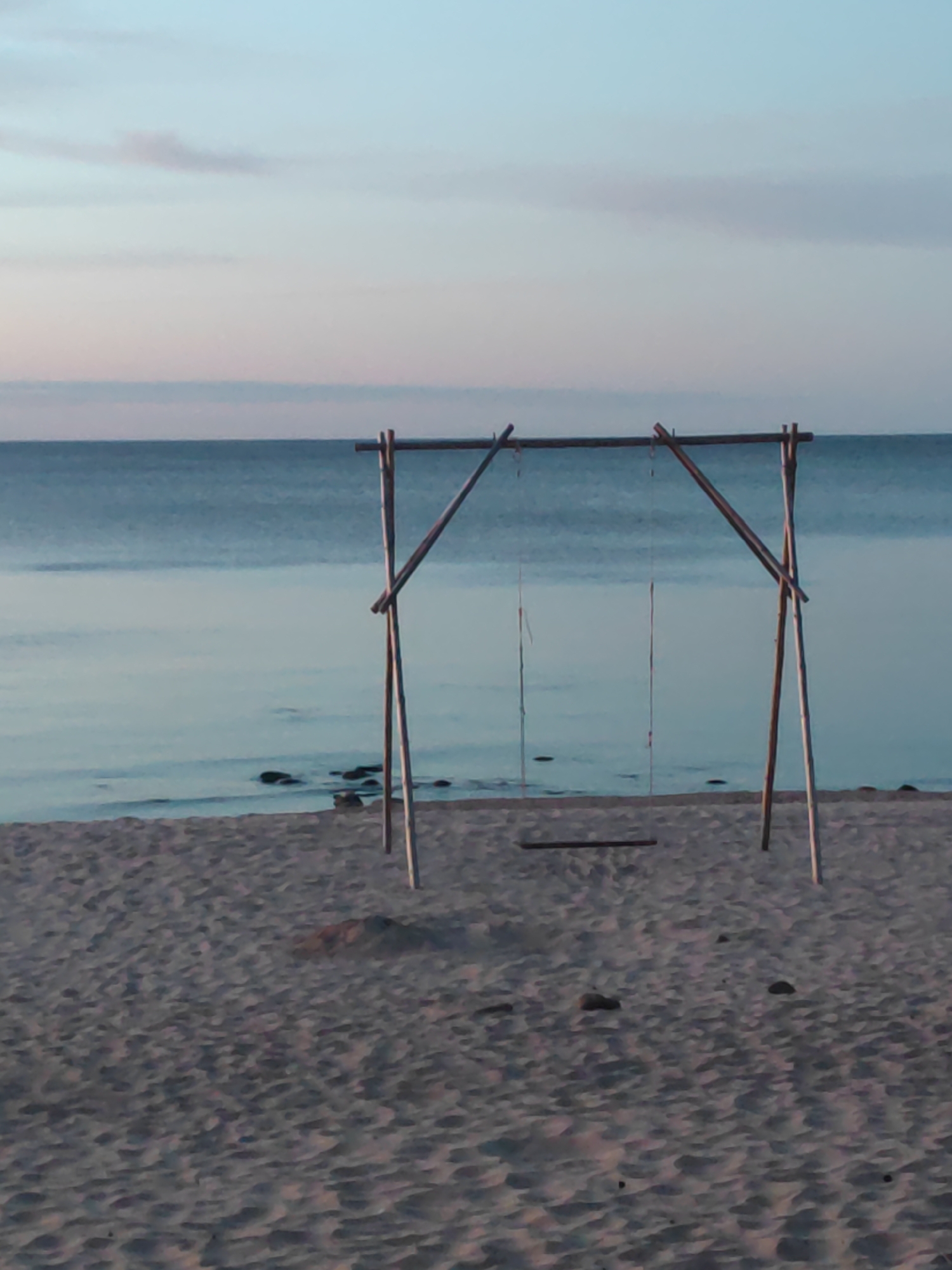 This is a daytime, eye-level, medium shot photograph of a beach and the sea. The scene is set on a sandy beach with a wooden swing set in the foreground. The swing set is constructed from bamboo poles. The sea is calm and blue, meeting a light blue sky with faint clouds. There are some rocks visible in the shallow water near the shore. The overall impression is one of tranquility and natural beauty. The location is Motornoe, Russia.