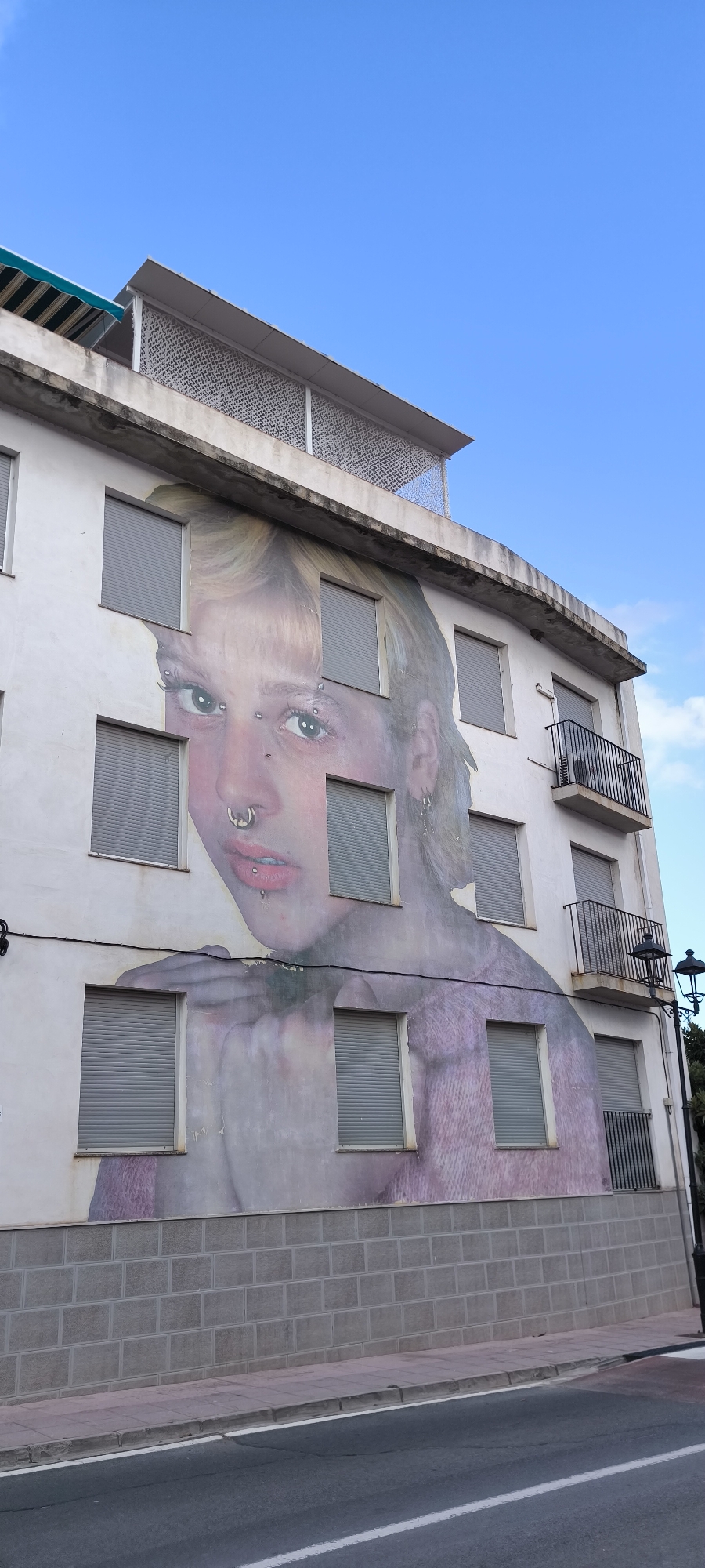 This is a full shot, taken from a low angle, of a building facade in Fanzara, Spain, covered in a large mural of a young woman's face. The mural is broken up by windows, creating a fragmented appearance. The woman has blonde hair, piercings on her nose and eyebrows, and is wearing a purple top. The building itself is white and has several windows with grey shutters and two balconies. The sky is blue with some clouds. A street lamp stands on the right side of the image. The bottom of the image shows a street and sidewalk.