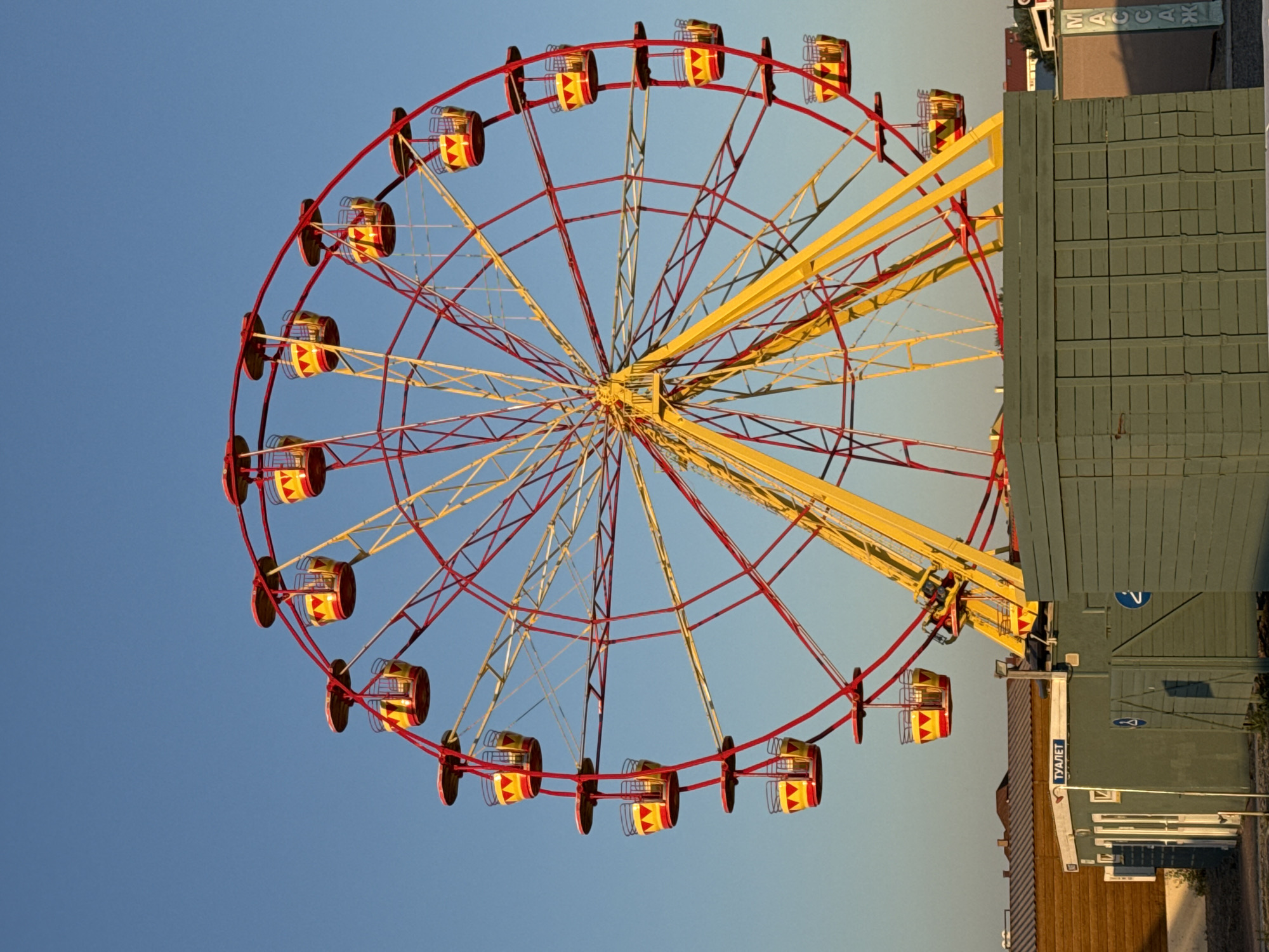 An outdoor shot captures a vibrant Ferris wheel against a clear, blue sky. The Ferris wheel is primarily red and yellow, with red support beams and a yellow central structure. It has numerous yellow and red gondolas, each with a striped pattern. At the base of the Ferris wheel, there are some low-lying buildings. One building, colored grey-green, has signs with blue symbols. To the left of this building, a smaller wooden structure is visible. The ground appears to be a mix of gravel and dirt. The overall scene suggests a recreational area, possibly an amusement park or a fairground, located in Novofedorovka, Ukraine.