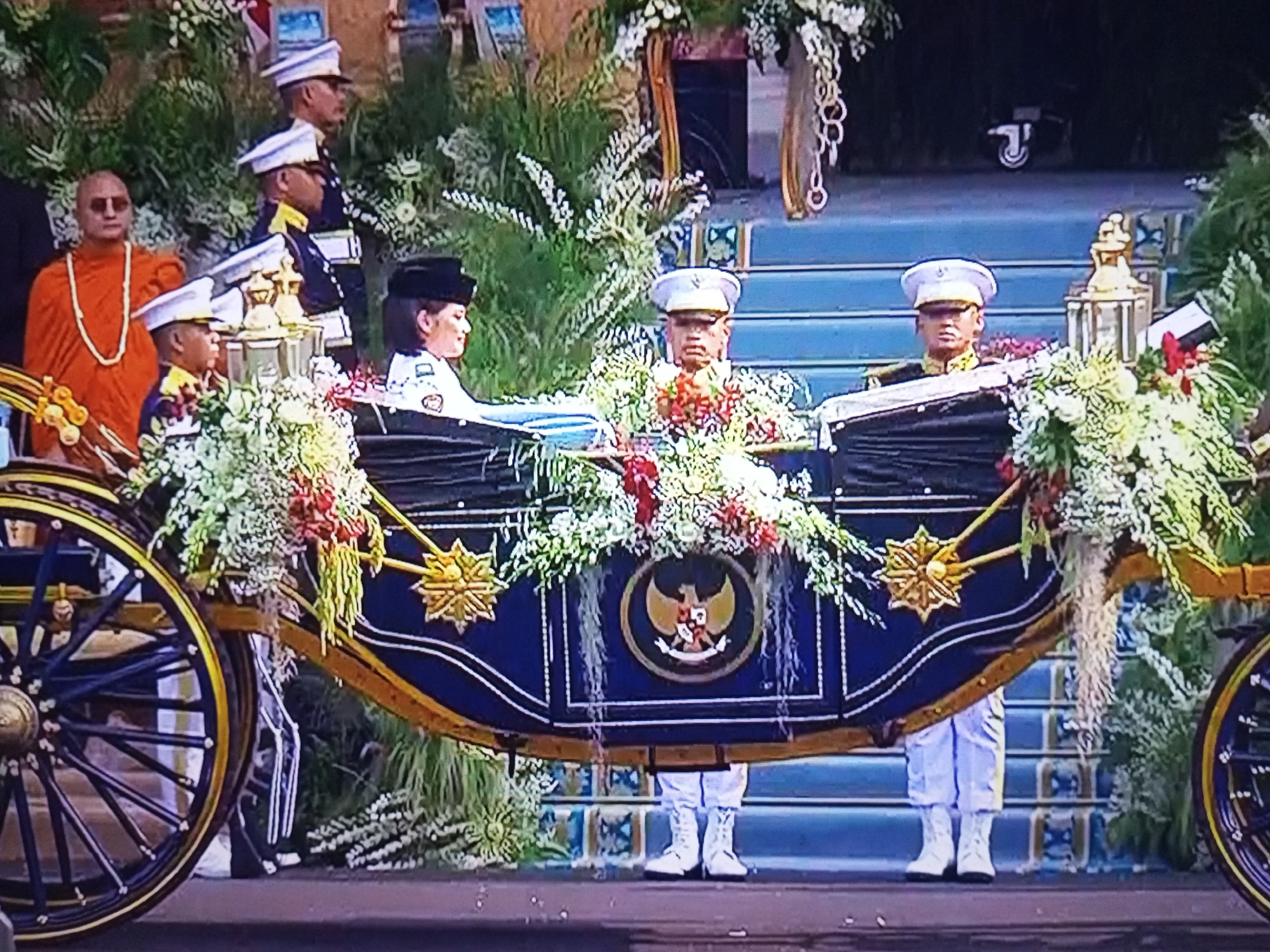 In the city of Banyudono, Indonesia, a solemn procession takes place. A horse-drawn carriage, adorned with abundant floral arrangements, serves as the central focus. Two individuals in ceremonial military attire, white hats and trousers, stand at attention within the carriage, appearing to be escorting it. To the left, a figure dressed in an orange robe and a white necklace, likely a religious leader, is partially visible, observing the proceedings. Behind the carriage, several other individuals in military uniforms, also wearing white hats, stand at attention. The carriage itself is elaborately decorated with a dark blue body featuring golden trim and a prominent national emblem in the center. The surrounding environment appears to be outdoors, with a backdrop of greenery and architectural elements suggesting a formal setting, possibly a parade or a state ceremony. The overall scene conveys a sense of respect and formality.