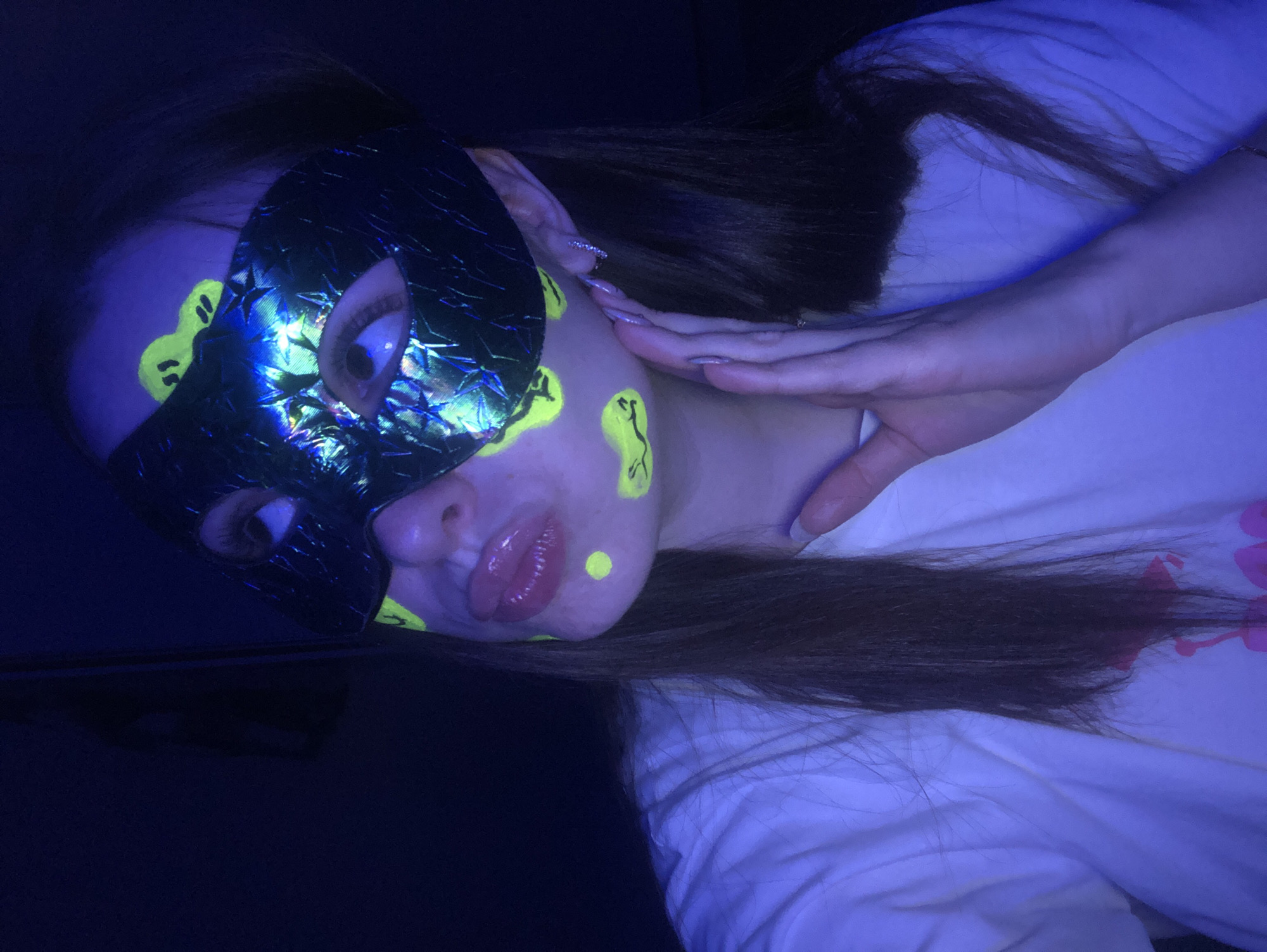 A woman wearing a black mask with iridescent reflections lies on her side on a white surface. The mask covers the upper part of her face, revealing one eye. She has yellow face paint in various shapes on her forehead, cheek, and chin. Her hair is brown and lies across the white surface, which appears to be a bed or similar soft surface. She is wearing a white top. Her hand is near her face, with visible fingernails. The lighting is dim with a blue hue, making the scene appear under a black light. The location is not identifiable as Noviy, Russia from the image.
