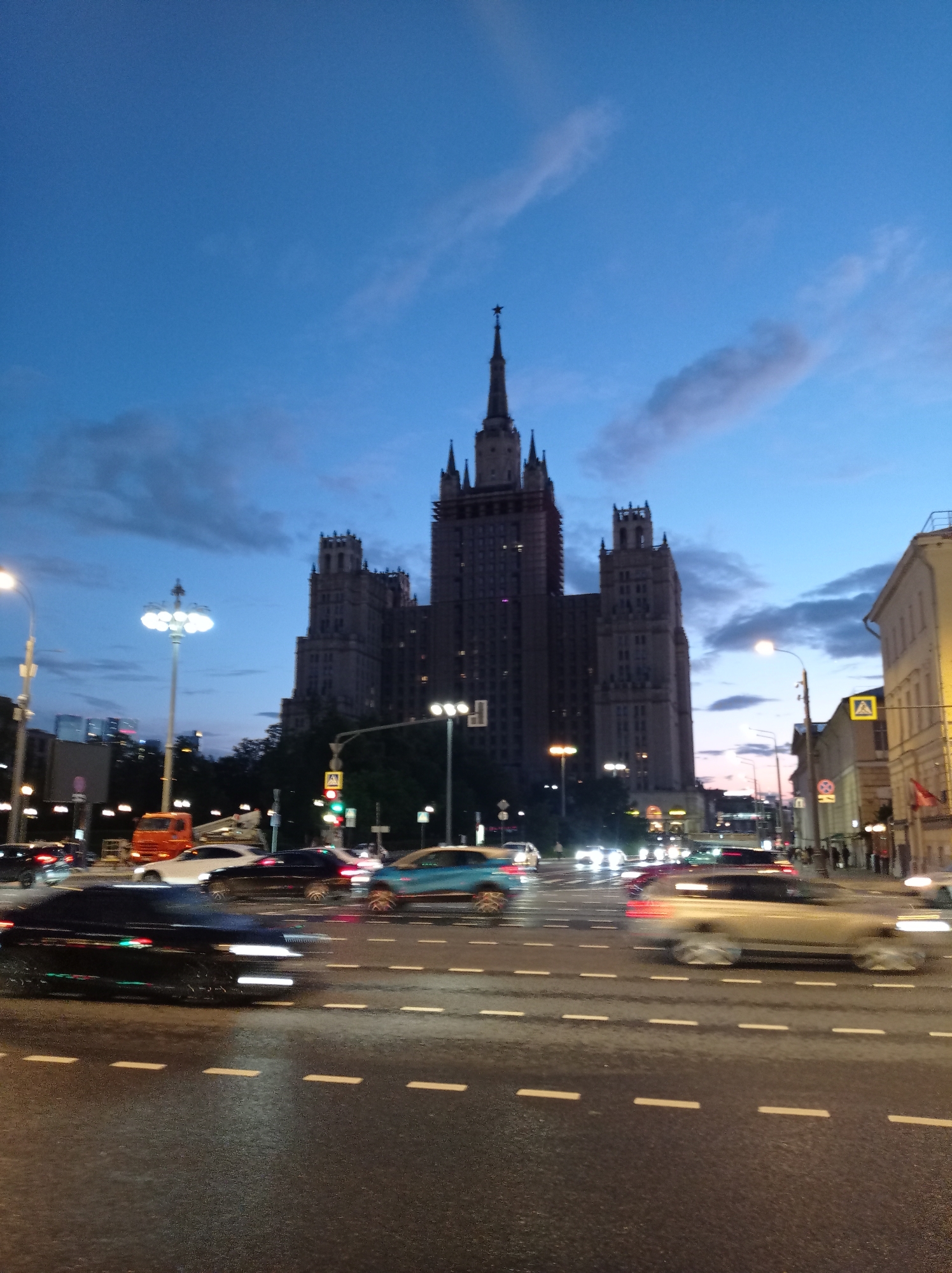 This is a photograph taken at dusk of the Kotelnicheskaya Embankment Building in Moscow, Russia. The iconic Stalinist skyscraper dominates the skyline. The foreground shows a busy street with cars in motion, their headlights and taillights creating streaks of light due to the long exposure. The road appears wet, reflecting the streetlights and the building's illuminated windows. Traffic lights are visible, some green, indicating ongoing traffic flow. Pedestrians are sparsely visible on the sidewalks. The sky is a deep blue with wispy clouds, suggesting the transition from day to night. The overall scene depicts a dynamic urban environment at twilight.