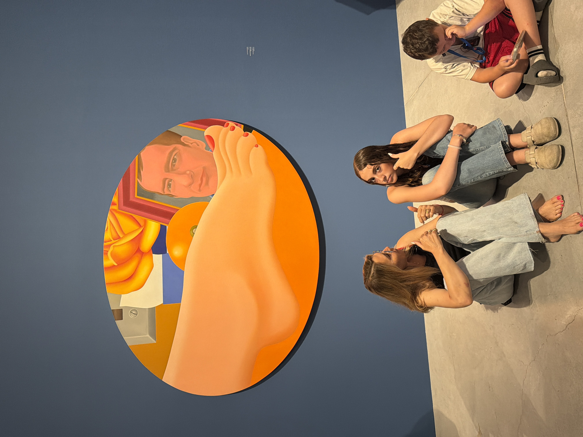 In the city of Tel-Aviv, Israel, three individuals are seated on a concrete floor in front of a large, oval-shaped artwork. The artwork, predominantly orange and red hues, depicts a close-up of a human foot and leg, an orange, a yellow rose, and a man's face within a framed section. To the left of the artwork, a woman with blonde hair, wearing a black tank top and light blue jeans, is seated and giving a thumbs-up gesture. Next to her, a young woman with dark, wet hair, wearing a sleeveless top and blue jeans, is also seated and giving a thumbs-up. To the right, a young boy in a white t-shirt and red shorts is seated and looking down at a phone or tablet. The wall behind the artwork is painted a dark blue.