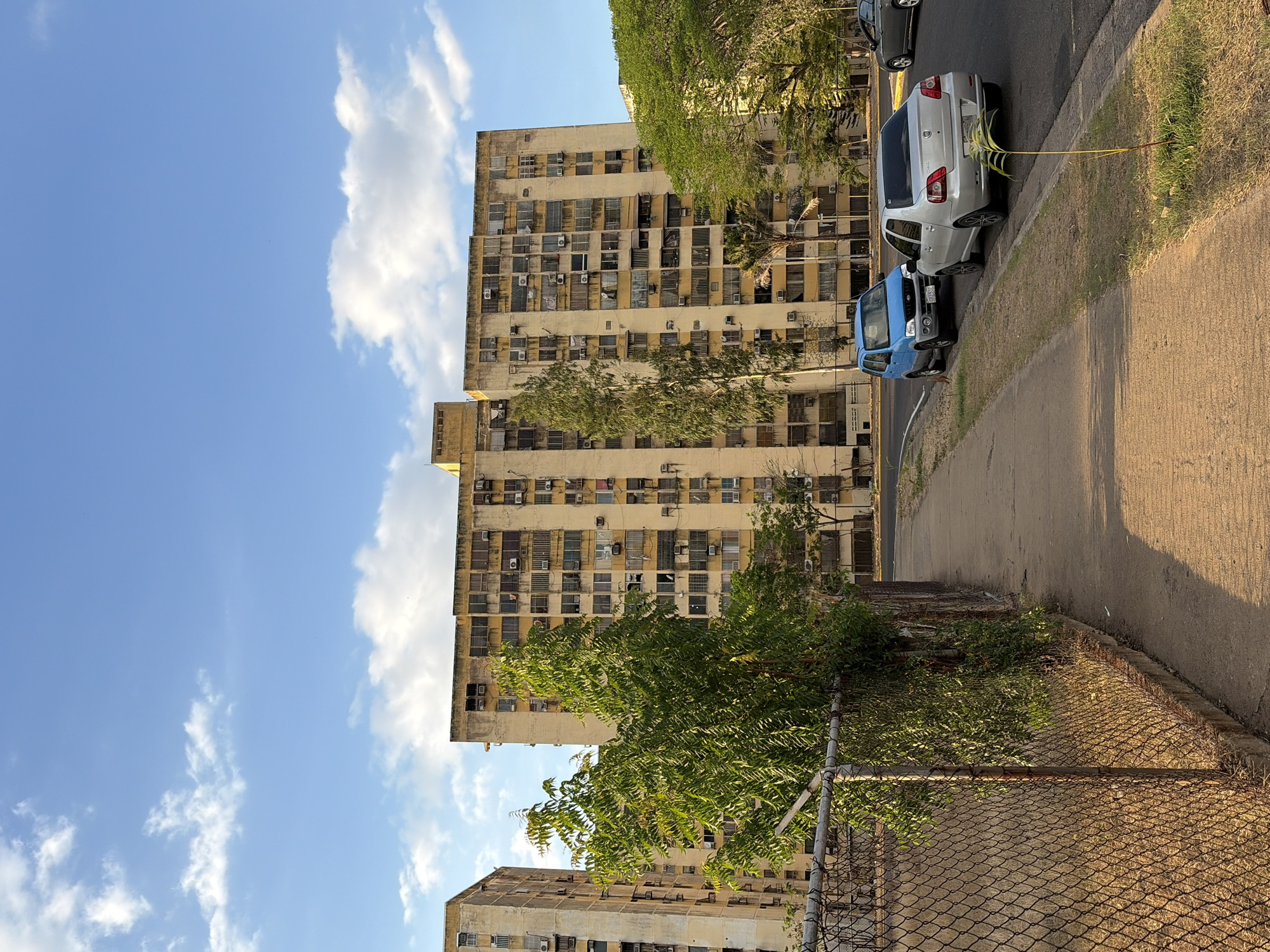 The image shows a street in Ciudad Guayana, Venezuela. A tall, aging apartment building is the primary focus, with numerous windows and what appear to be air conditioning units. Parked along the street are a blue vehicle and a silver car. A chain-link fence and some greenery are in the foreground. The sky is blue with scattered clouds. The road surface is paved, and there is a grassy area beside the road.
