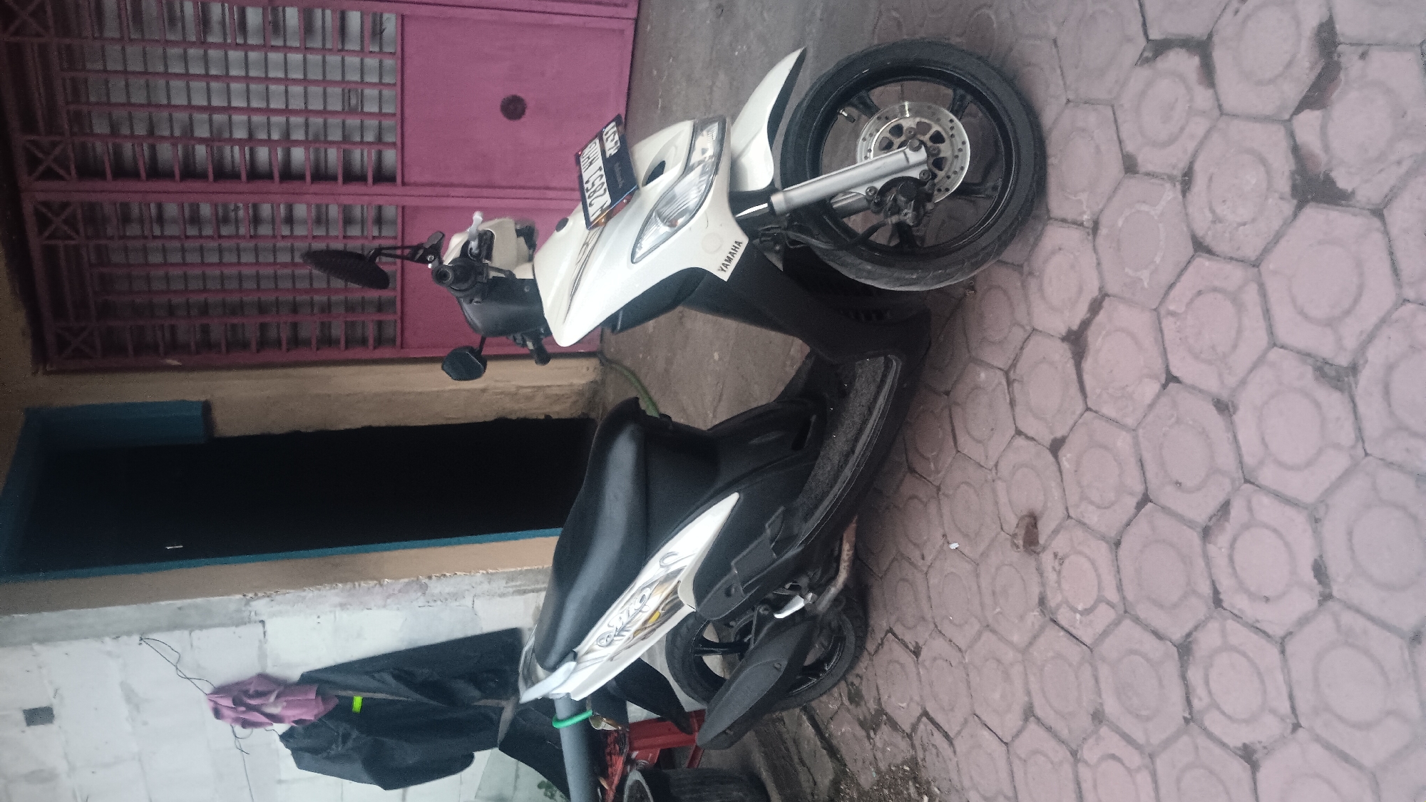 A white Yamaha scooter is parked on a patterned concrete ground in front of a building with a pink metal door. To the left of the scooter, a black jacket and a pink cloth are hanging on a wire attached to the building's white wall. The building appears to be a residential or commercial structure, with a dark doorway to the left of the pink door. The ground is made of hexagonal concrete tiles. The scene is likely outdoors. The license plate on the scooter reads "B 2853 NHD".