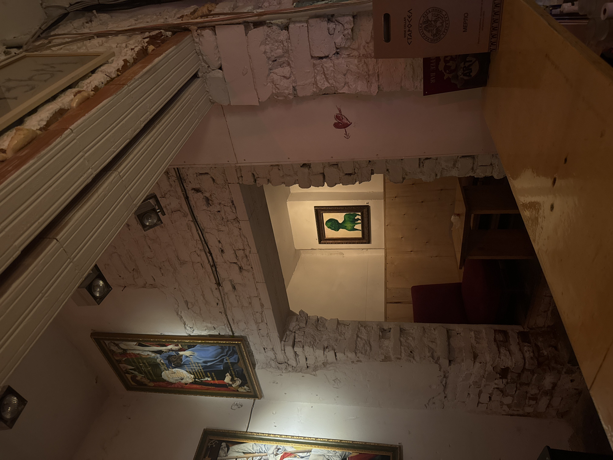 This image captures the unique interior of a building in Sofia, Bulgaria, likely a cafe, bar, or restaurant, characterized by a rustic and eclectic design. The scene is illuminated by artificial indoor lighting, suggesting an evening or nighttime setting.
The space features prominent exposed brick walls with rough plasterwork, giving a textured, unfinished appearance. Multiple framed paintings are visible; a large one on the left wall depicts a religious scene, possibly Madonna and child, while a smaller one through an archway in the background shows a distinctive green, abstract figure. Small spotlights are integrated into the ceiling and walls, providing focused illumination.
Architecturally, the space appears to be multi-level, with visible arches and openings revealing different sections of the interior. A prominent light-colored wooden surface, possibly a bar or a large table, occupies the right foreground. In the background, a red upholstered chair or sofa is partially visible. Exposed building materials, such as wood beams and some insulation foam, contribute to the raw, industrial-chic aesthetic.
Notable details include a small, stylized red heart with an arrow painted on an upper wall. Visible text includes "WINE CELLAR", "СТАРОСЕЛ" (Starosel), "МЕРЛО" (Merlot), and "COFFEE BAR" printed on a cardboard box located near the top right. Another small sign clearly reads "ART". No individuals are present, allowing the focus to remain on the architectural details and decor.