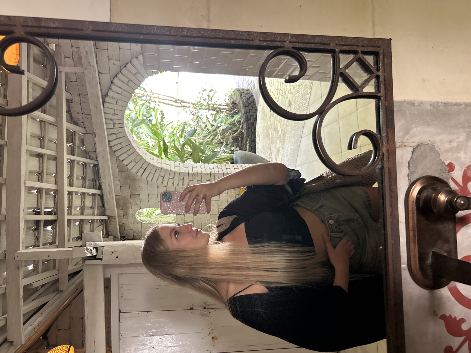 A person is visible in the reflection of a mirror. The person is holding a phone and appears to be taking a selfie. The environment reflected in the mirror shows a building structure with white walls, an arched opening leading to a garden area with plants, and a tiled or brick-like construction. The mirror is framed with a decorative ironwork design. The location is Kuta Selatan, Indonesia.