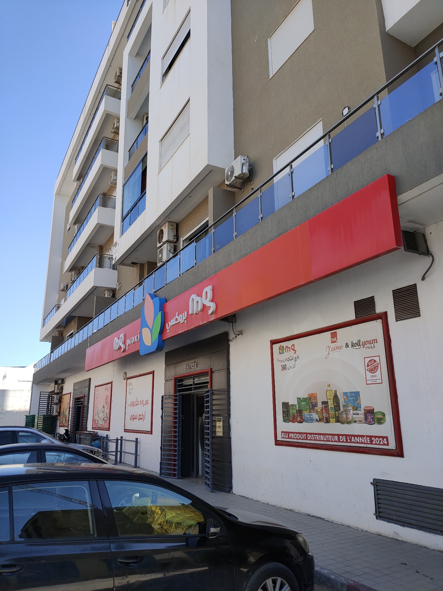 A low-angle exterior shot captures a modern, multi-story building with a white facade and a brown upper section, under a clear blue sky. The building features multiple balconies with glass railings. At the street level, a bright red awning stretches across the building, and below it, a store with a prominent "MG proxi" logo is visible. The store's entrance is open, revealing metal security gates. To the right of the entrance, a large advertisement poster is displayed on the wall, showcasing various grocery products and text that reads "ELU PRODUIT DISTRIBUTEUR DE L'ANNÉE 2024." A black car is parked in the foreground, partially obscuring the sidewalk and street. Several other cars and a motorcycle are parked further back. The overall scene suggests a commercial area in a city, likely in Tunisia, given the Arabic text on the signage.