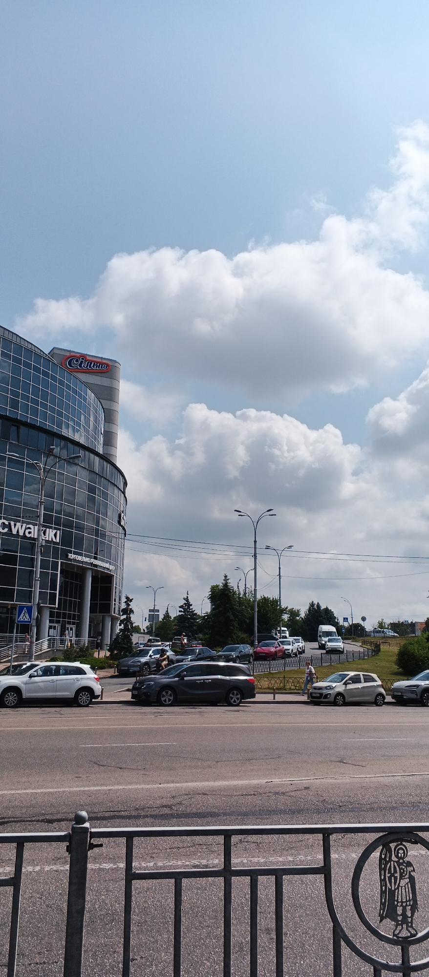 This is a vertical, eye-level shot of a modern building with a curved glass facade in Kyiv, Ukraine. The building, labeled "Silpo" in red lettering on a grey pillar, appears to be a supermarket or shopping center, as indicated by the text "ТОРГОВЕЛЬНО-РОЗВАЖАЛЬНИЙ ЦЕНТР" (Trade and Entertainment Center) below the main entrance. In front of the building, a street runs horizontally across the frame, with several cars parked along the curb and some in motion.
The sky is a bright blue with scattered white, fluffy clouds. A black metal fence with a circular cutout featuring an angel is visible in the foreground, partially obscuring the street and sidewalk. Several streetlights and traffic signals are present along the street. The overall impression is an urban scene on a clear, sunny day.