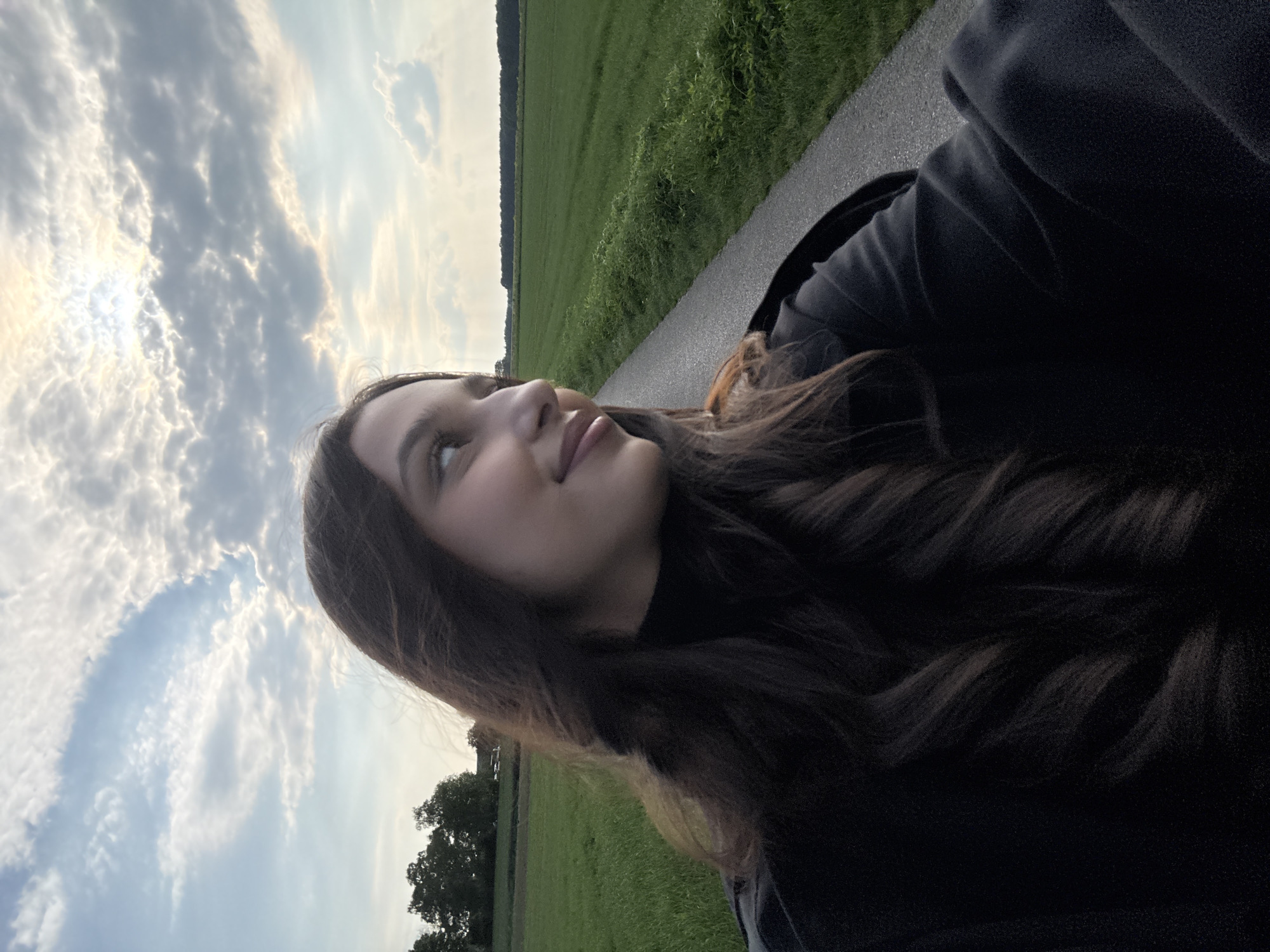 A young woman stands on a paved road in a rural area in Wegberg, Germany. She is wearing a black turtleneck and a dark, possibly black, jacket or coat. She is turned slightly to the left and looking upwards towards the sky. The sky is filled with clouds and the sun is visible. A large grassy field extends into the distance on either side of the road, with trees visible along the horizon.