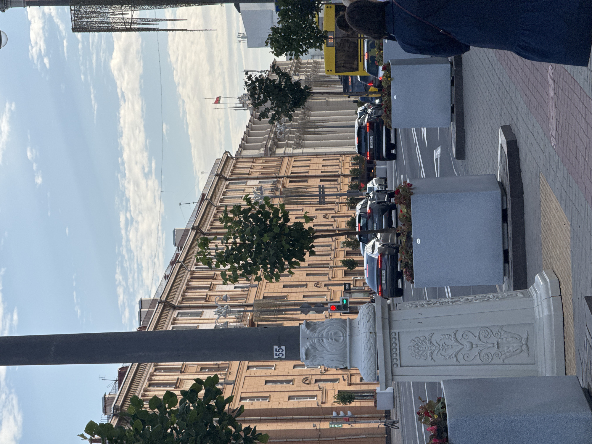 The image depicts a street scene in Minsk, Belarus, likely on Independence Avenue, given the architectural style of the buildings and the presence of a yellow bus. In the foreground, a large concrete planter contains red and pink flowers. To its left is a tall, decorative lamp post with a number "55" visible on it. The street is lined with cars in motion, including a dark-colored Audi with its brake lights illuminated. On the right side of the street, another large concrete planter is visible, and a woman in a dark blue dress is walking away from the camera. The background features a long, multi-story building with a neoclassical facade, characterized by numerous columns and arched windows. A red and green traffic light is visible, indicating controlled traffic flow. The sky above is partly cloudy with a light blue hue. The overall impression is a bustling urban environment.