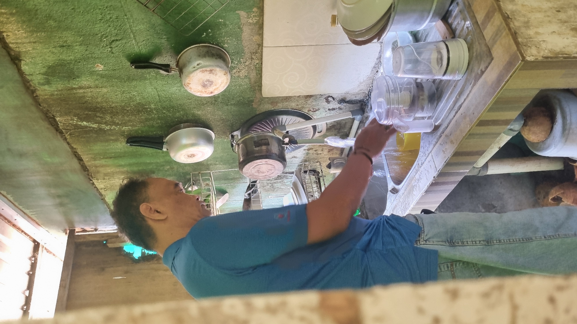 A man wearing a blue collared shirt and light-wash jeans is standing at a kitchen sink and washing dishes. The sink is made of stainless steel and contains yellow dish soap and water. A faucet is on, with water running into the sink. There are various kitchen items on the counter and hanging on the wall, including pots, pans, dishes, and containers. The walls appear to be made of concrete and are painted green, with visible water stains and some mildew. The kitchen appears to be located in an outdoor or semi-outdoor area, with a tin roof visible at the top of the frame. The overall impression is one of a functional, rustic kitchen in a rural or informal setting. This scene is likely from Birobuli Utara, Indonesia, a residential area known for its informal housing and local businesses.