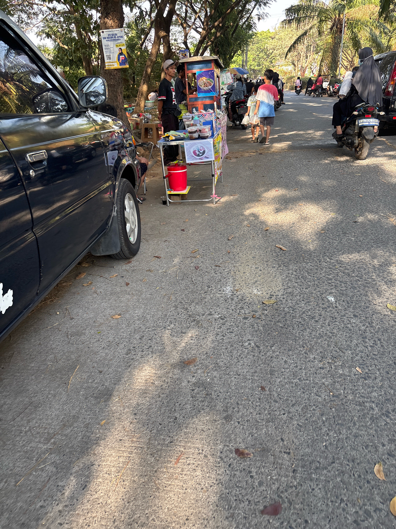 A sunny, outdoor scene on a paved road in Telagamurni, Indonesia, depicts a roadside vending area. A dark blue SUV is parked on the left, its side and wheel visible. In the middle ground, a food cart with a colorful display of food items and condiments is set up. A man in a dark t-shirt and a baseball cap stands behind the cart, attending to customers. Several other people are visible, some on motorcycles, and two children walking away from the cart. A woman wearing a hijab rides a motorcycle in the background. Palm trees and other foliage line the road, with a poster advertising an event attached to a tree. The ground is asphalt, with fallen leaves scattered across it, and dappled sunlight creates shadows.
