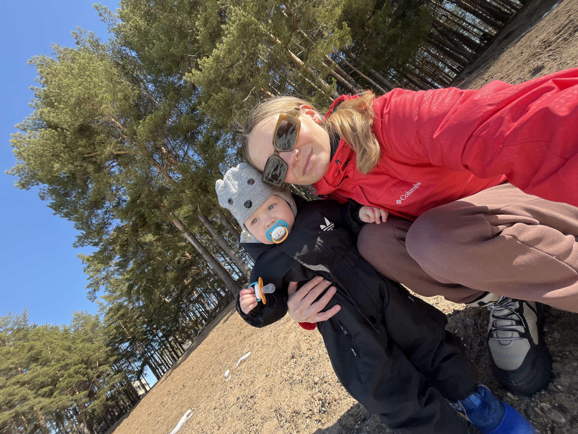 The image shows a woman and a baby outdoors, likely in a park or forested area, as evidenced by the presence of tall trees in the background. The woman is wearing a red jacket and sunglasses, and she has light-colored hair. She is holding the baby, who is wearing a black snowsuit and a grey hat with bear ears. The baby has a pacifier in its mouth and is holding another pacifier in its hand. They are positioned on a slightly angled surface that appears to be covered with light-colored dirt or sand. There are patches of snow visible in the lower part of the image. The woman is wearing brown sweatpants and gray and black hiking shoes, while the baby is wearing blue boots. The location is likely in Chaikovsky, Russia.