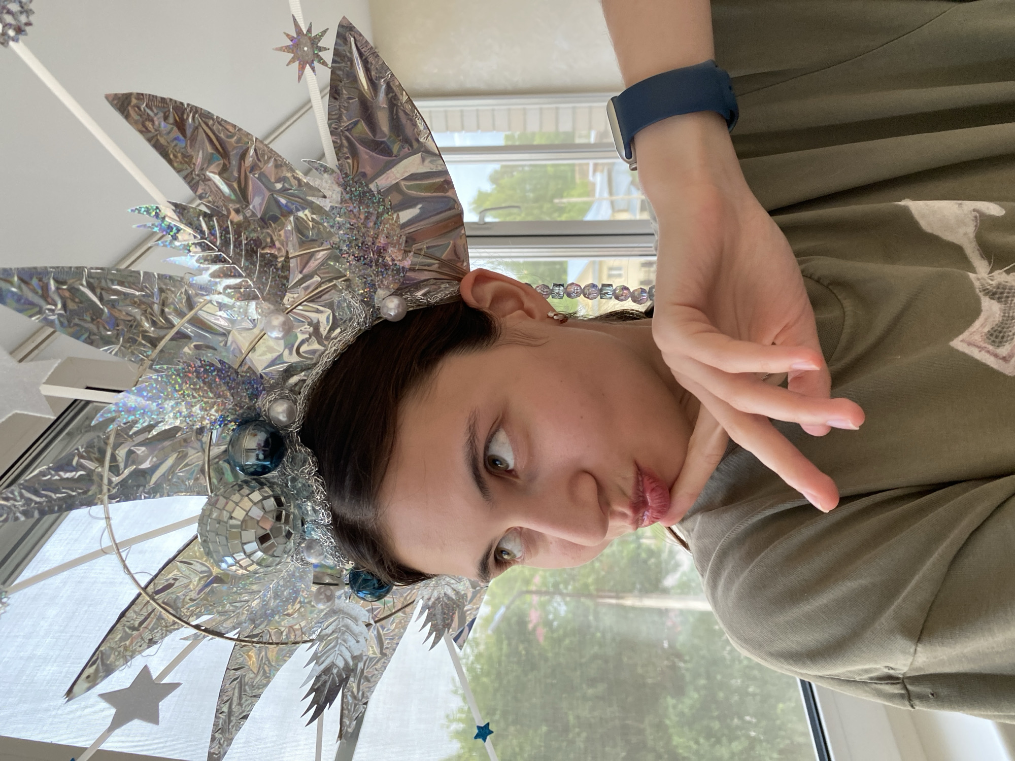 A young woman stands indoors, posing with a large, elaborate, silver headdress adorned with disco balls, blue ornaments, and pearl-like beads. She is wearing a green t-shirt and a dark blue smartwatch on her left wrist. Her hand is placed under her chin, and she is puckering her lips, looking upwards and to her left. The background shows a window with a blurred view of trees and buildings, suggesting an urban or suburban setting. The lighting indicates it is daytime. The headdress is the central and most prominent object in the image, overshadowing other details. No specific landmarks or context are provided to place this image in Votkinsk, Russia.